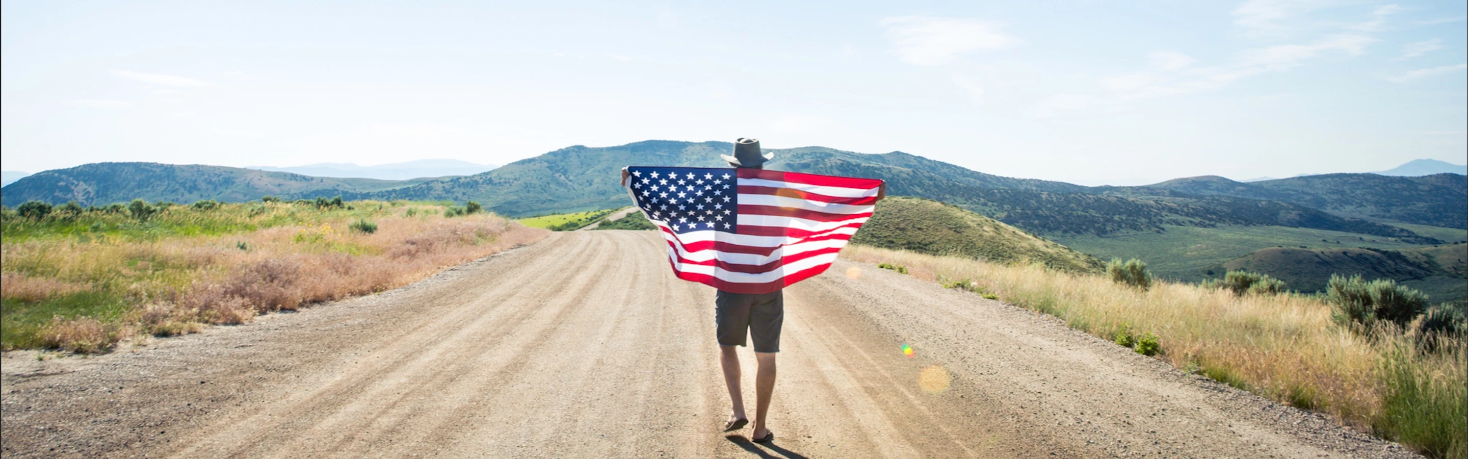 Person walking down a dirt road holding an American flag behind their back with outstretched arms.