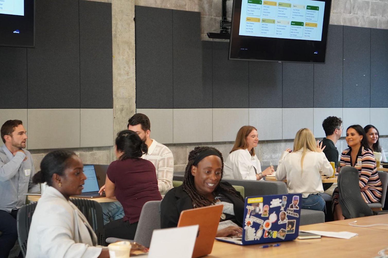 Group of people working on laptops and collaborating at various tables in an open room with large tv monitors hanging from the wall. 