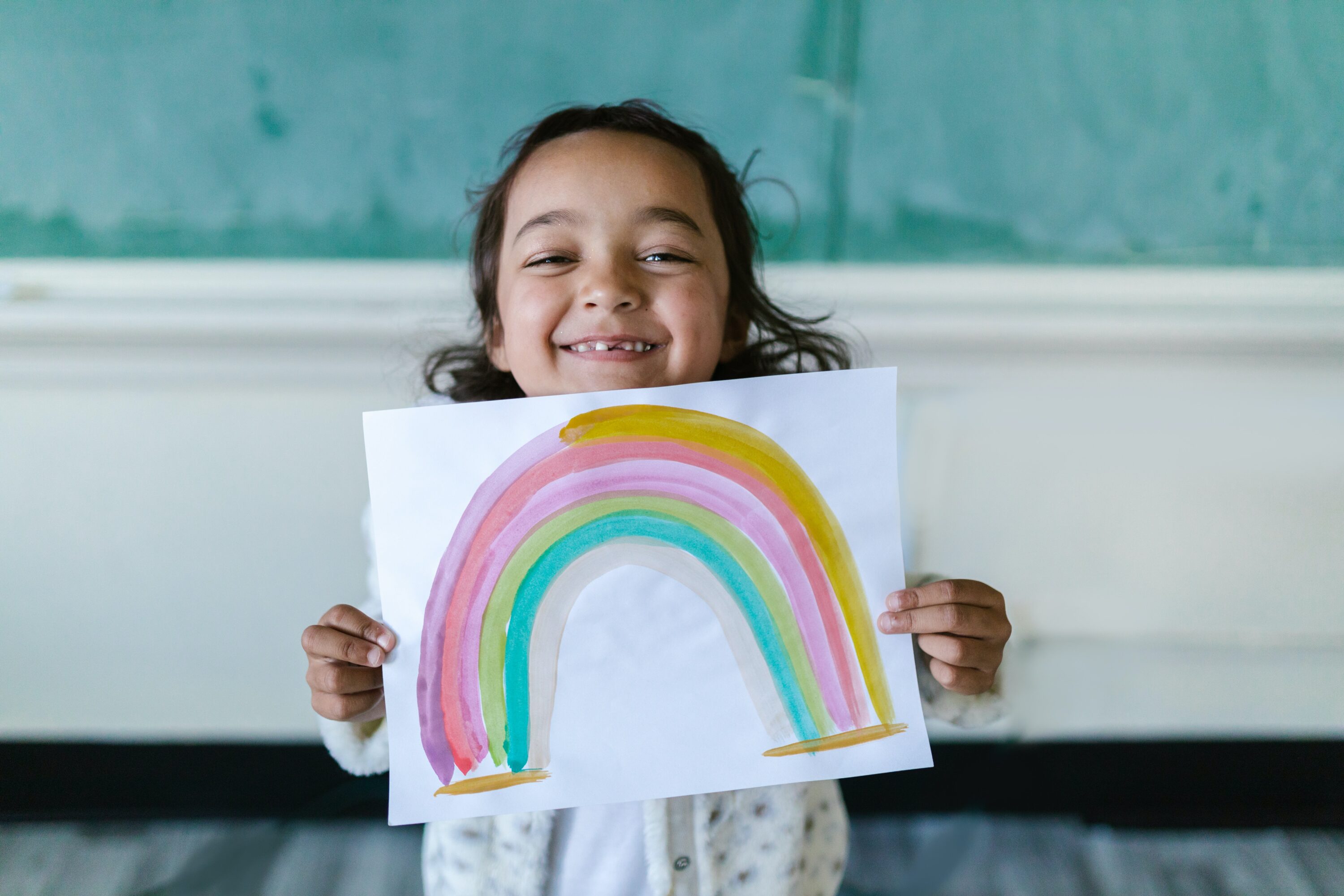 little girl holding white paper with rainbow drawing