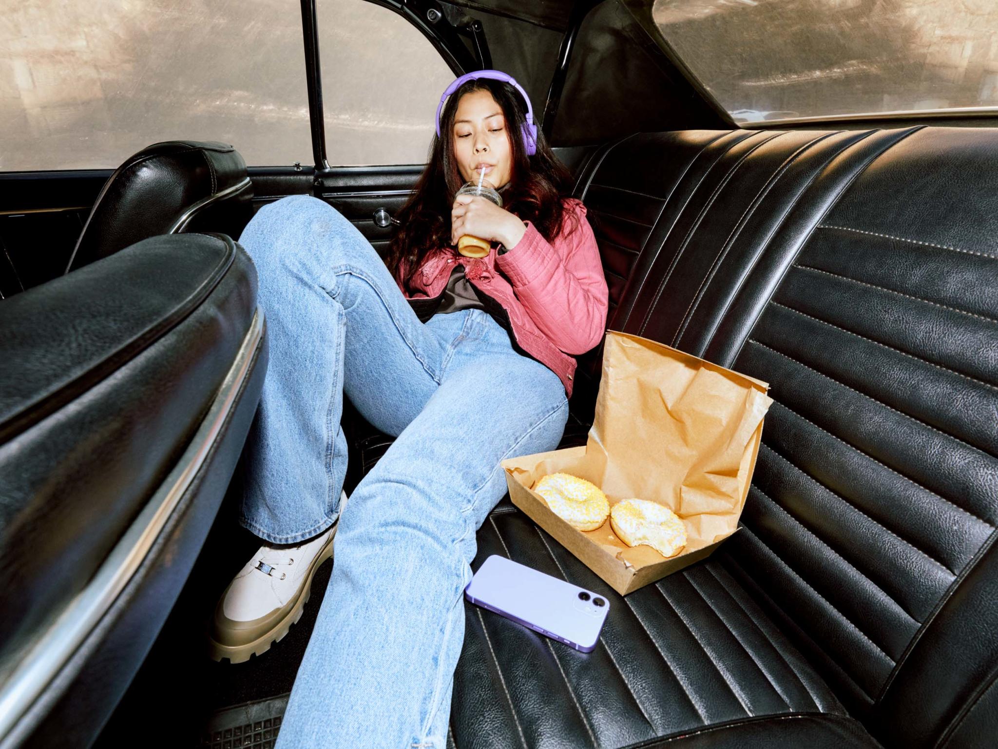 Młoda dziewczyna siedzi na tylnym siedzeniu samochodu i słycha audiobooka popijając napój przez słomkę.