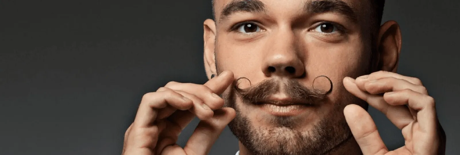 Pessoa com bigode estilo guidão, a segurar o rosto com ambas as mãos sobre fundo cinzento escuro