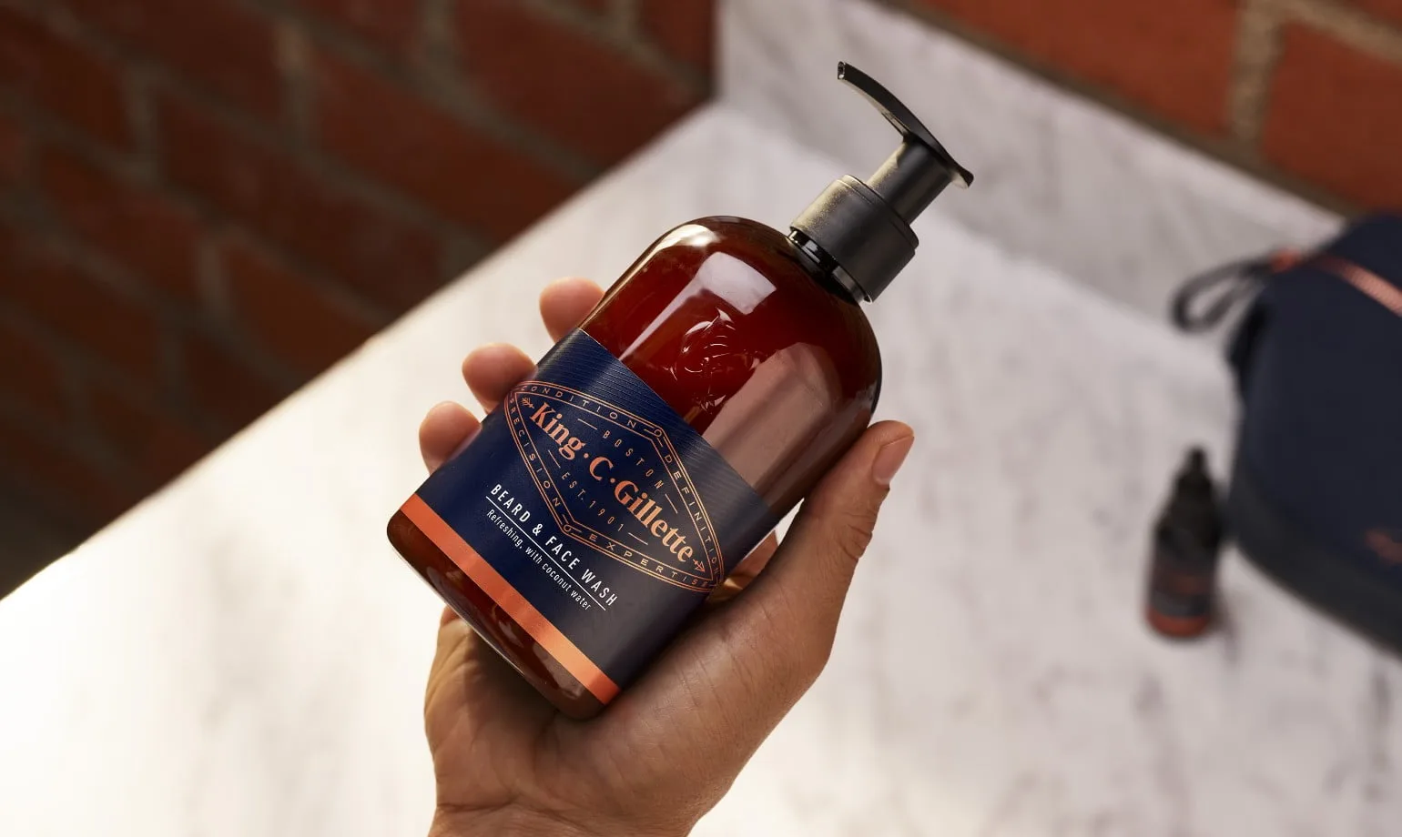 Mão segurando um frasco de Gel de Limpeza para Barba e Rosto King C. Gillette com rótulo azul e cobre