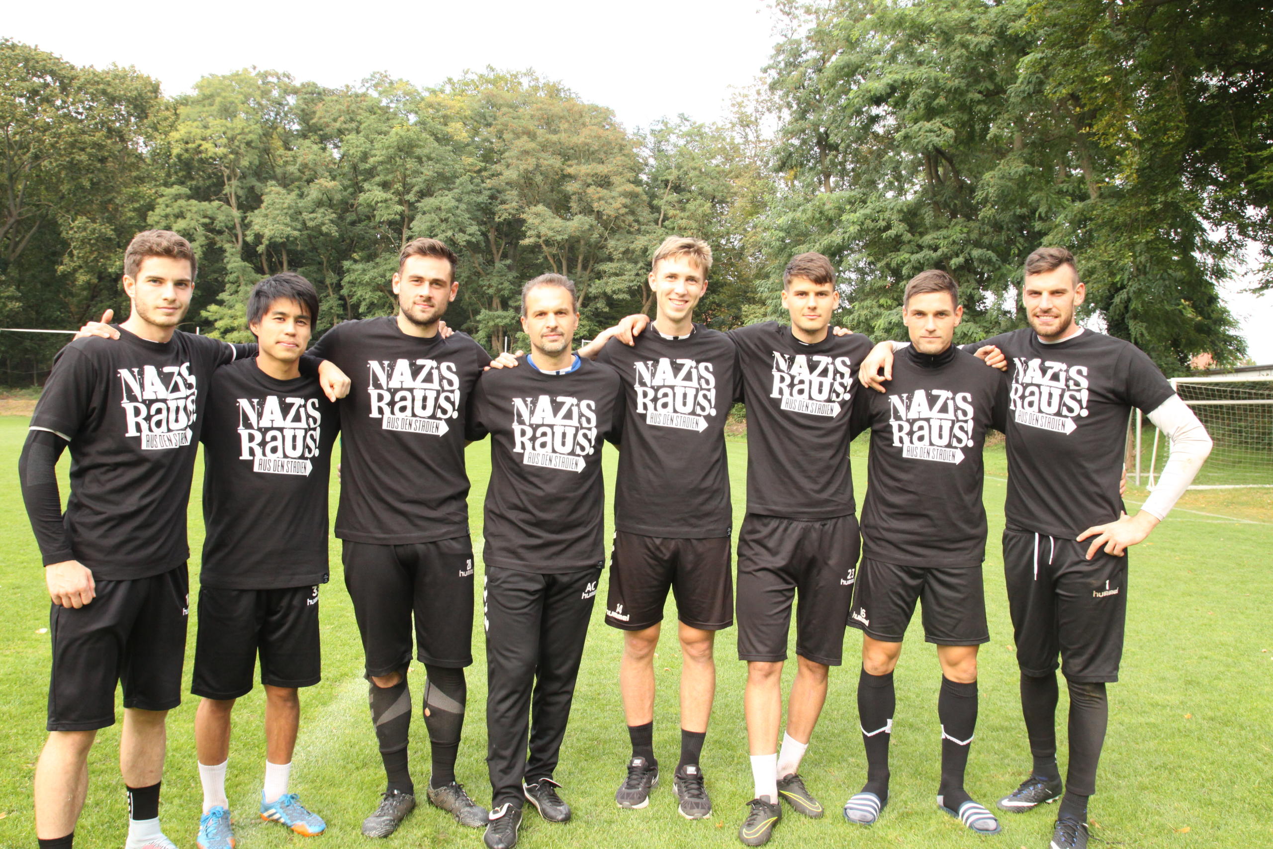 Acht sportlich gekleidete Männer stehen Arm in Arm auf einem Fußballfeld. Alle tragen ein schwarzes T-Shirt mit dem "Nazis raus aus den Stadien"-Logo aufgedruckt.