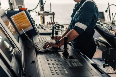 Photo of a person in a boiler suit on the bridge of a ship, standing up and typing on a laptop with one hand. 