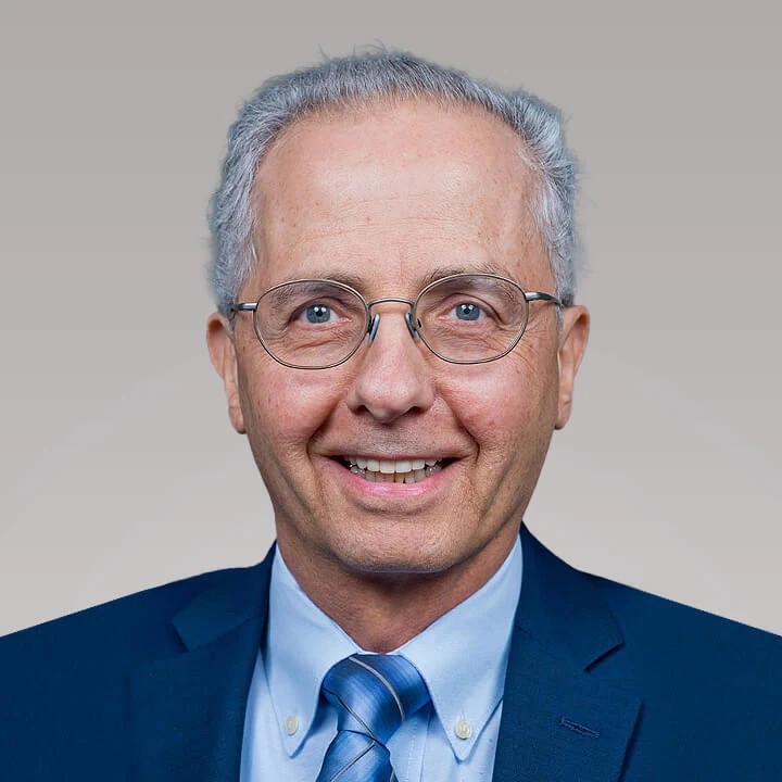 Headshot image of Gary Campenella. The image background is a light taupe color. Gary is wearing silver oval glasses, a dark royal blue blazer, a light blue button-up shirt, and a gradient blue tie. 