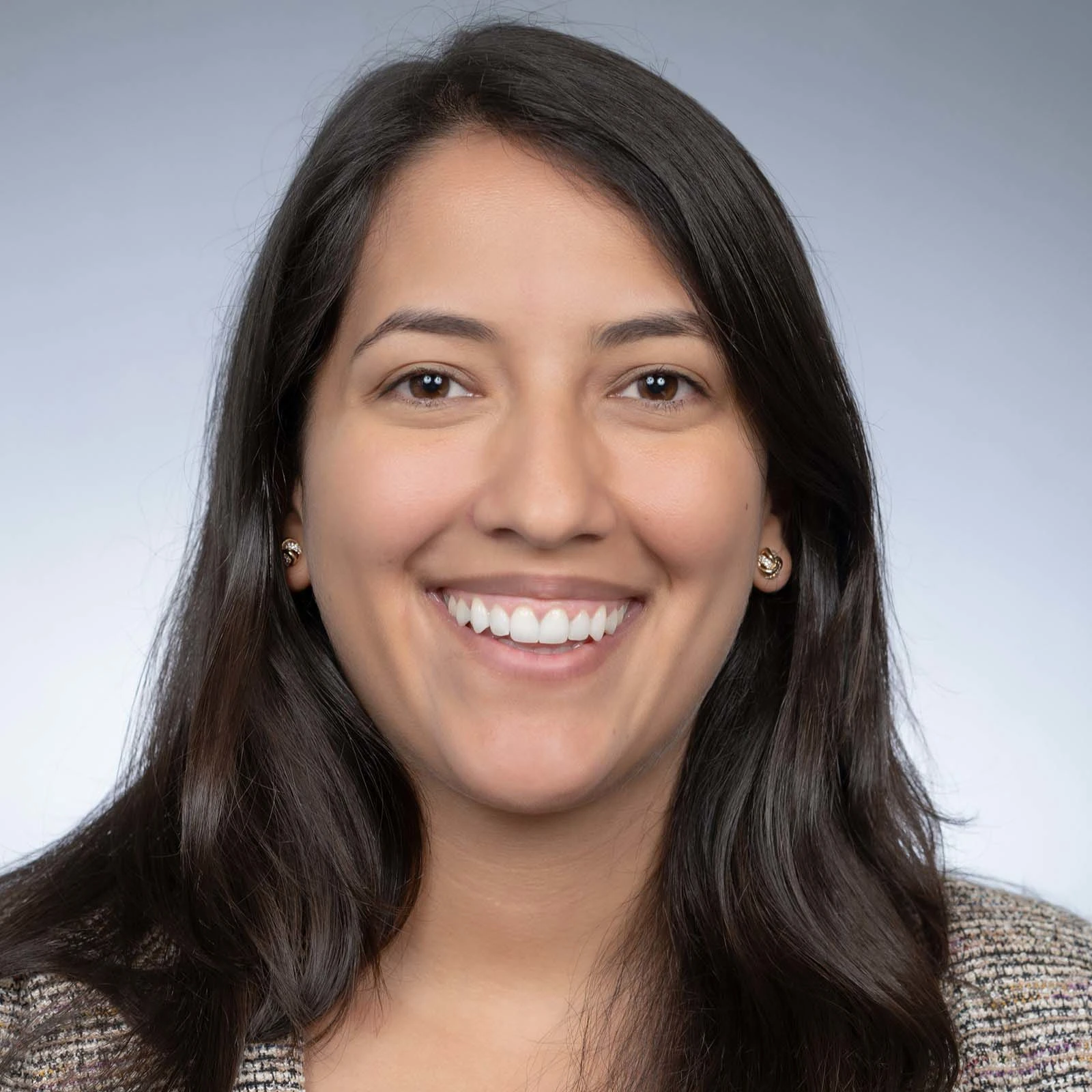 Headshot image of Melanie Cappel. The medium grey background fades into a light grey color. Melanie is smiling and wearing small gold earrings and a beige twill blazer. 