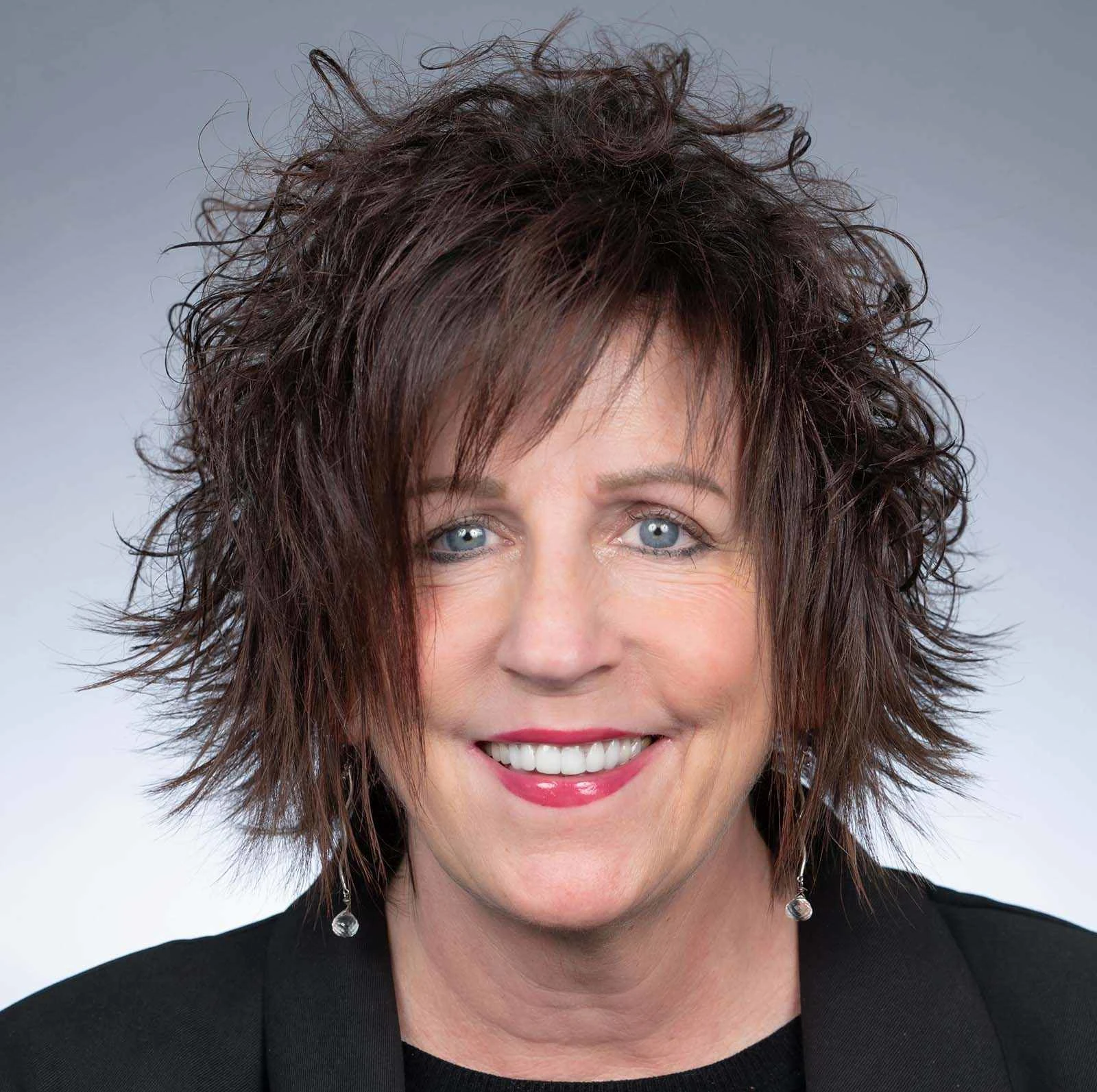 Headshot of Director of PACEpartners Operational Excellence, Lori Frank. The medium grey background fades to a lighter grey. Lori is smiling and wearing a black boat neck shirt and a black blazer. 