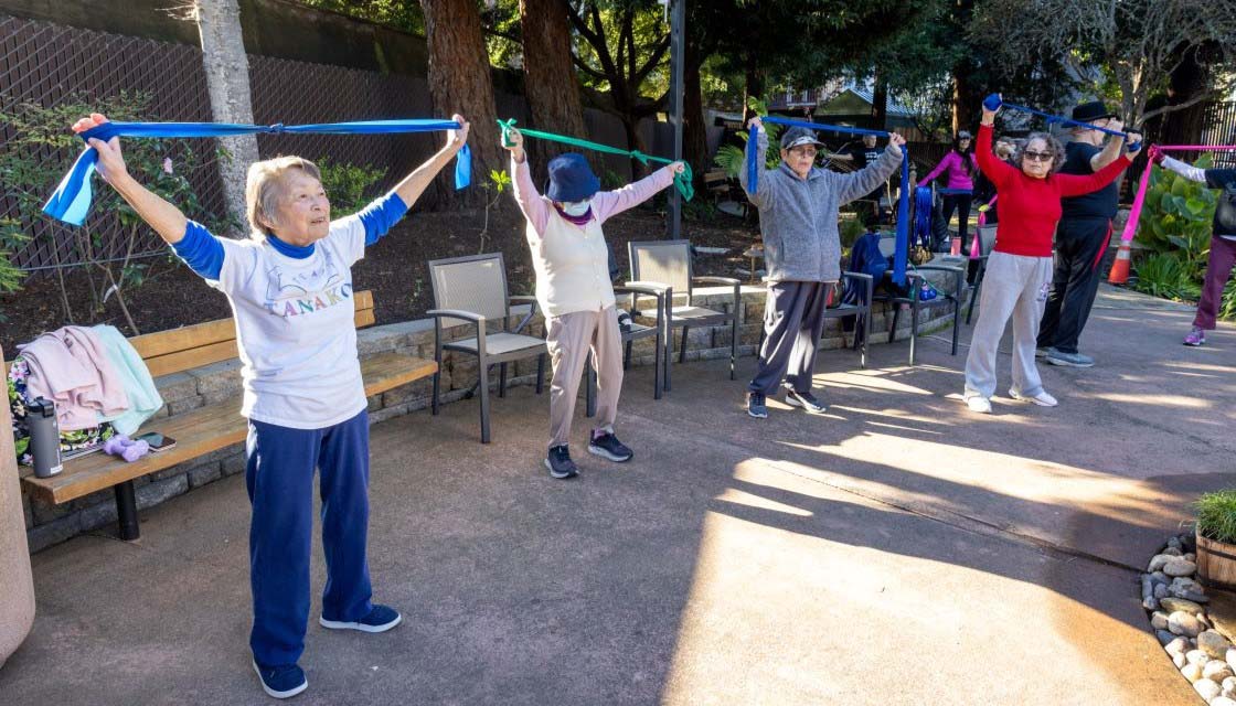 Image of a body dynamics class that took place at the On Lok 30th Street Senior Center Garden area. The image shows 5 seniors stretching their arms above their heads with stretching ribbon. 