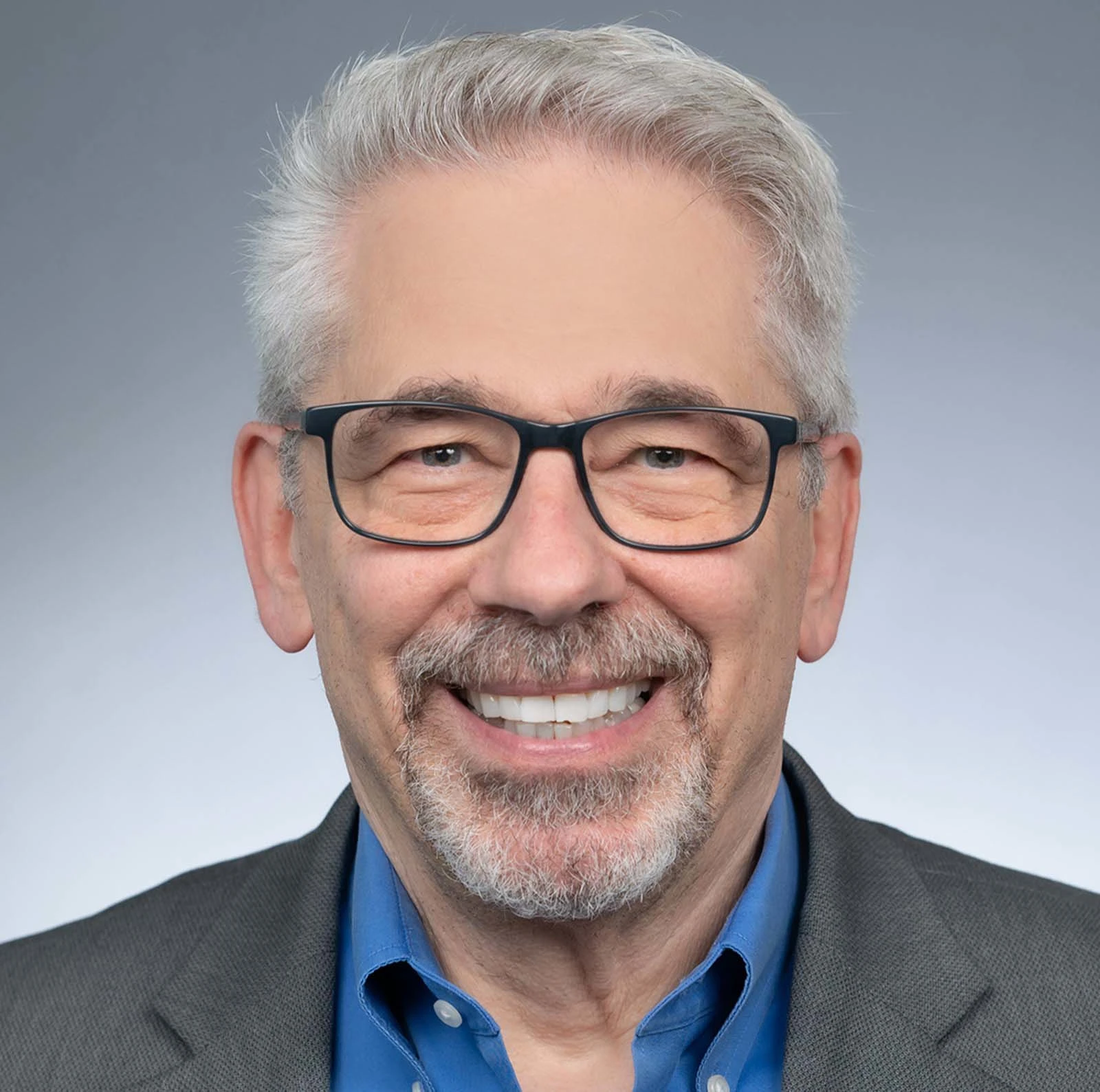 Headshot of the PACEpartners team lead, Sandy Rodgers. The medium grey background fades to a lighter grey. Sandy is wearing black framed glasses, a royal blue button-up shirt, and a dark grey blazer. 