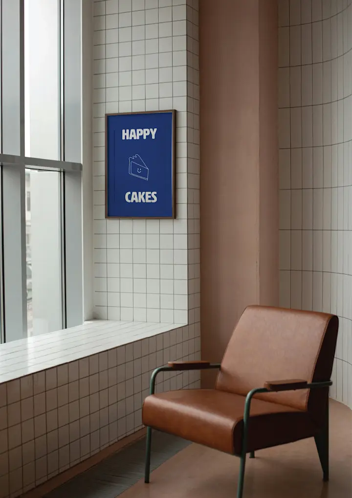 Happy Cakes Bakery Aesthetic: This Image Is A Branding Mockup For "Happy Cakes," Showcasing How The Brand's Identity Would Appear In A Physical Space, Likely Within A Coffee Shop Or Bakery Setting. The Image Features A Modern, Minimalist Interior Space With A Warm And Cozy Atmosphere. A Brown Leather Armchair With Metal Armrests Is Positioned In The Foreground, Suggesting A Comfortable Seating Area Where Customers Can Relax. The Chair’s Clean Lines And Industrial Design Elements, Such As The Metal Frame, Align With The Brand’s Modern Aesthetic. The Walls Are Tiled With White, Grid Like Ceramic Tiles That Add A Subtle, Yet Industrial, Texture To The Room. This Choice Of Wall Covering Enhances The Modern And Slightly Utilitarian Feel Of The Space, Which Contrasts Nicely With The Warmth Of The Leather Chair. On The Wall Next To A Large Window Is A Framed Poster Displaying The "Happy Cakes" Logo. The Poster Is A Deep Blue Color, Providing A Strong Contrast Against The White Tiles. The Logo Consists Of The Brand Name "HAPPY CAKES" Written In Bold, White Uppercase Letters, With The Smiling Cake Slice Positioned Between The Words.