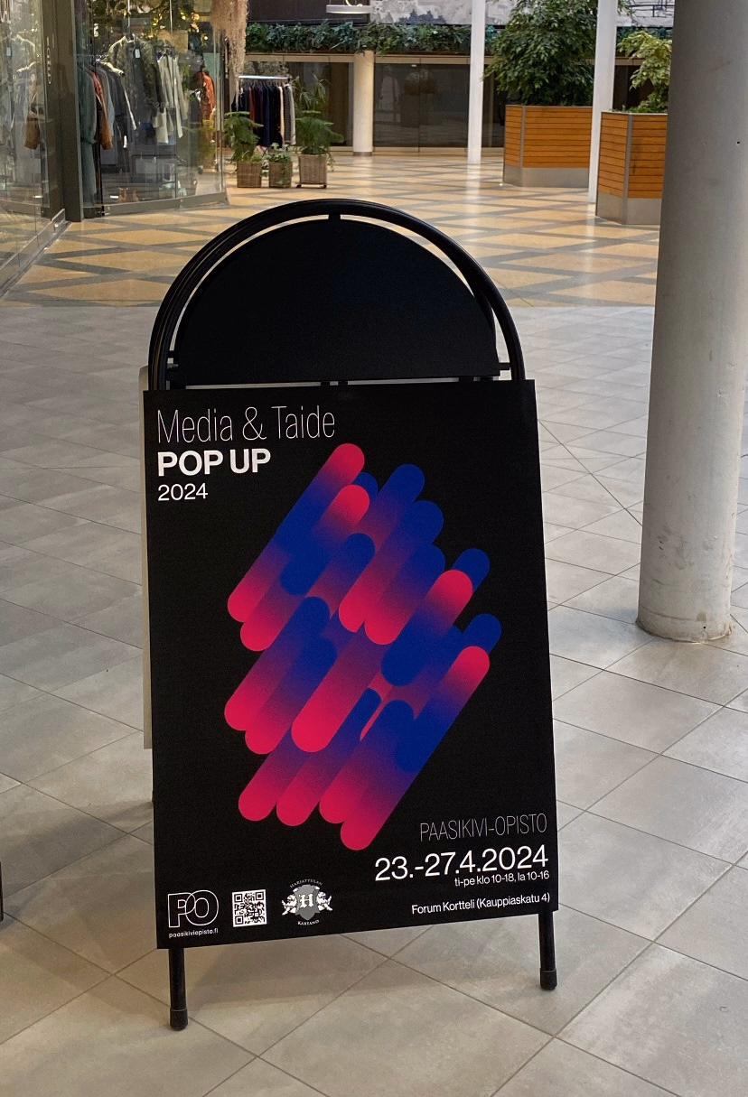 A-ständijuliste ostoskeskuksessa, teksti 'Media & Taide Pop Up 2024', sinipunaisin liukuvärimuodoin.