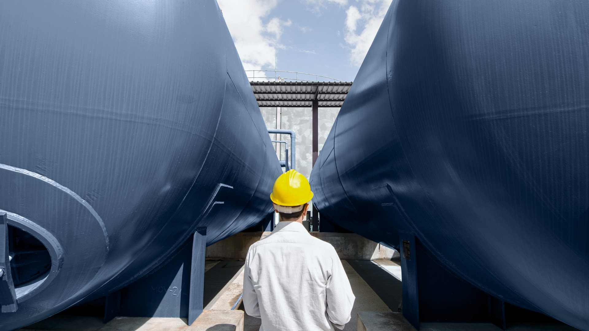 Mitarbeitender mit Schutzhelm inspiziert große industrielle Speichertanks in einer Energieanlage.