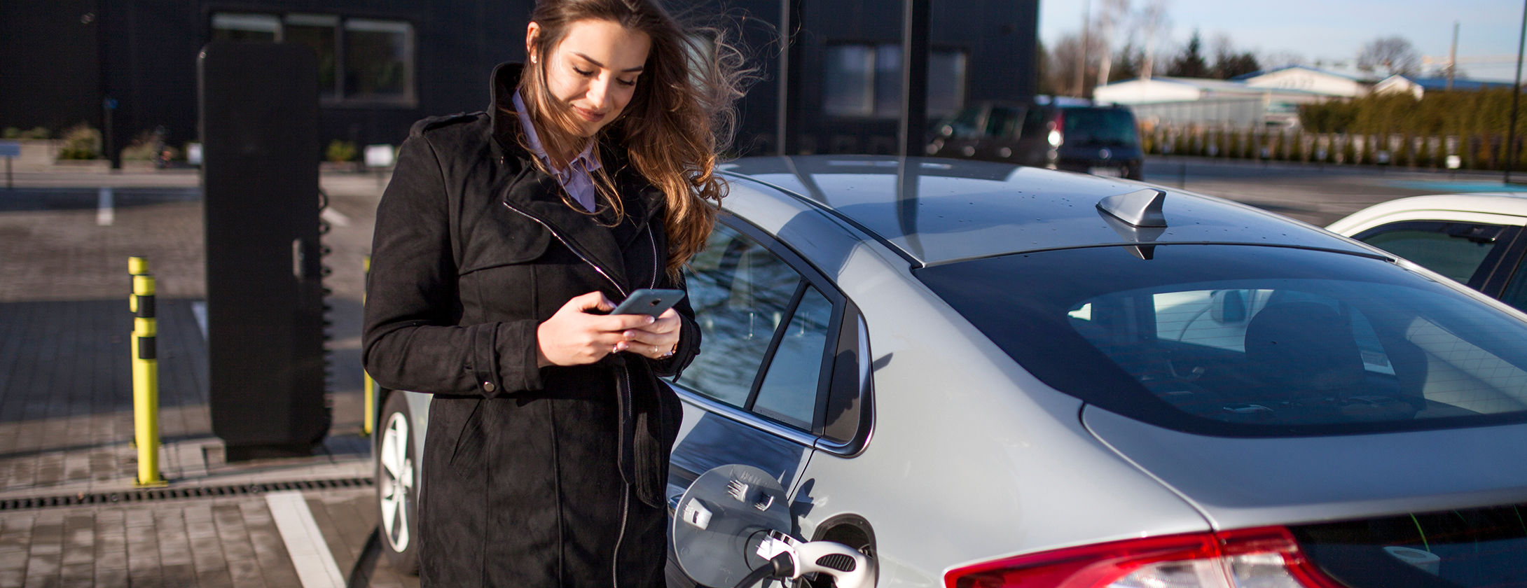 Una donna usa il suo cellulare fuori dalla sua auto elettrica mentre questa viene ricaricata in un parcheggio