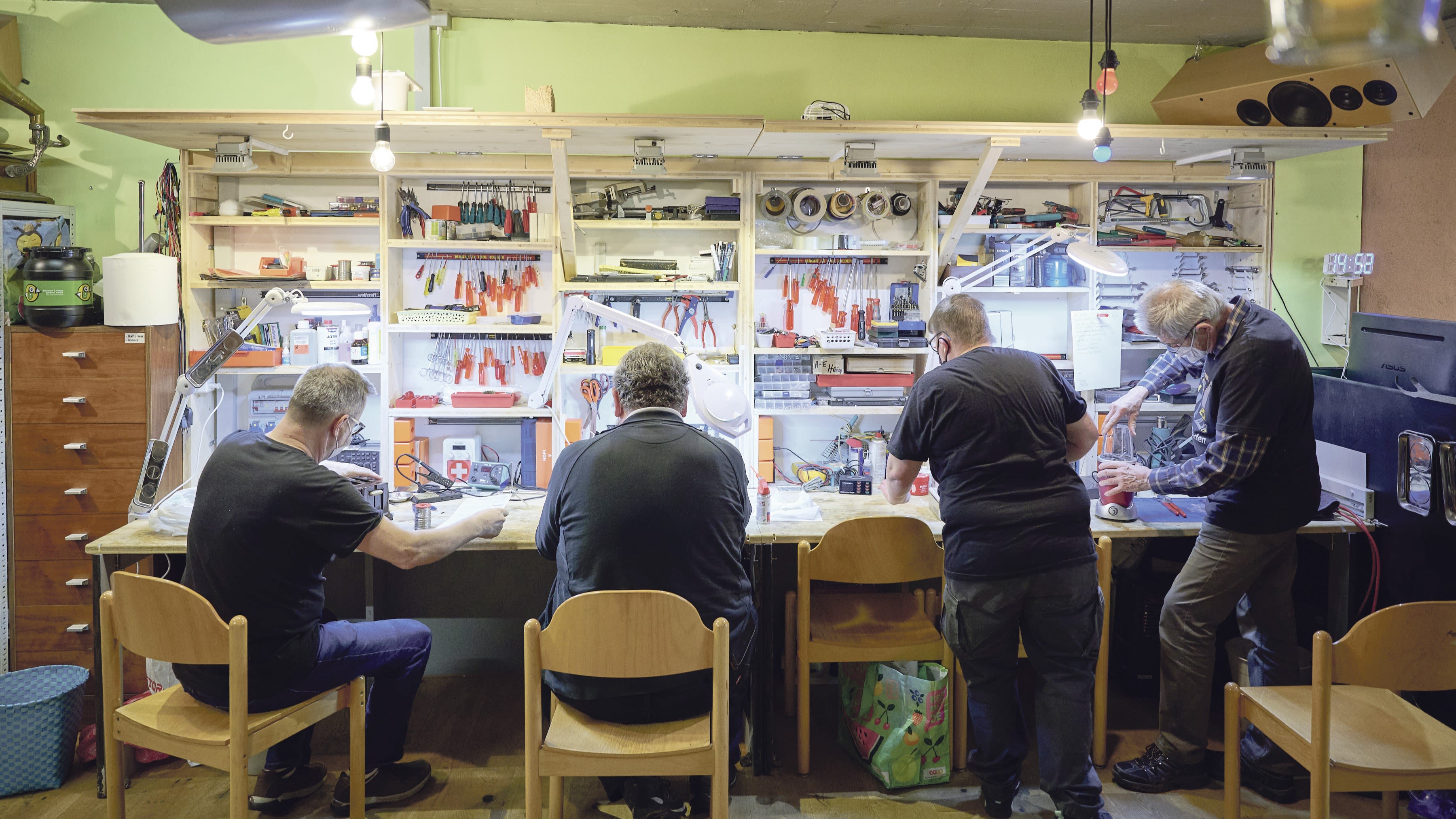 Vier Männer sitzen in einer Werkstatt und reparieren kaputte Elektrogeräte.