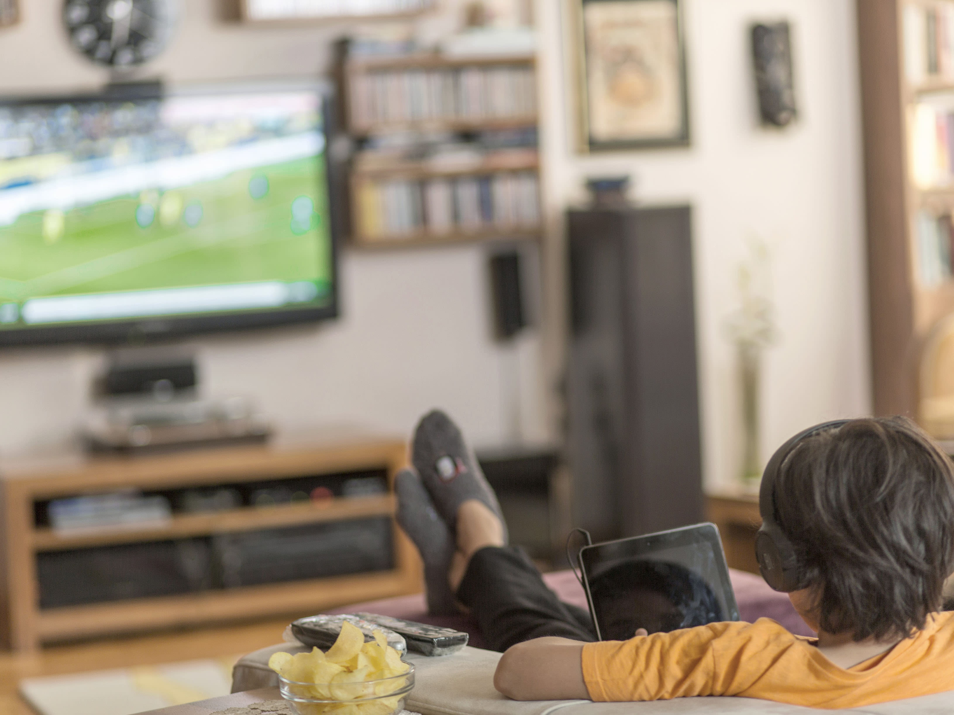 Junge mit Kopfhörern liegt auf dem Sofa und schaut Fussball im Fernseher.