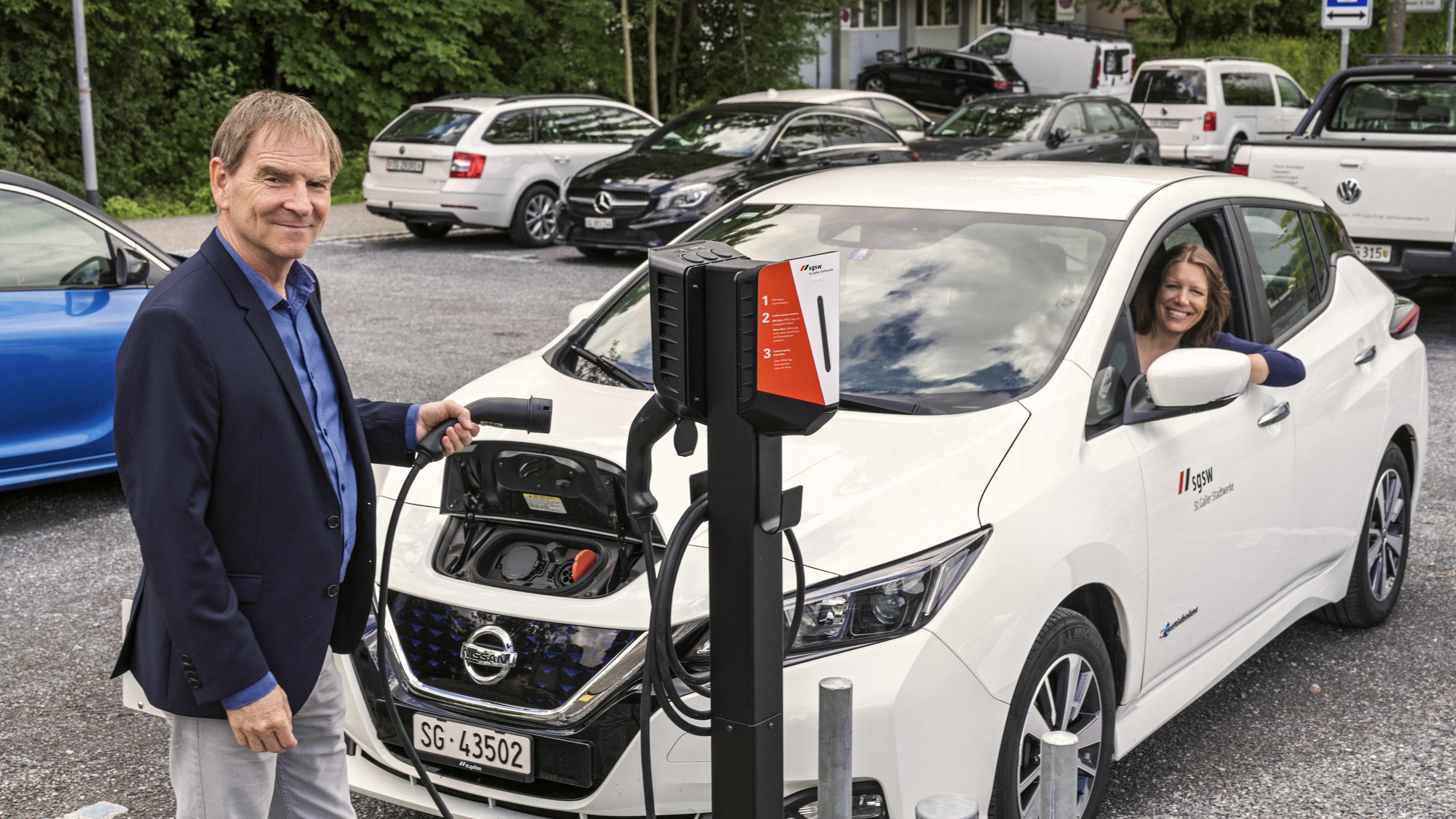Un homme connecte la voiture électrique à la borne de recharge. Une femme est assise dans la voiture et regarde par la fenêtre ouverte.