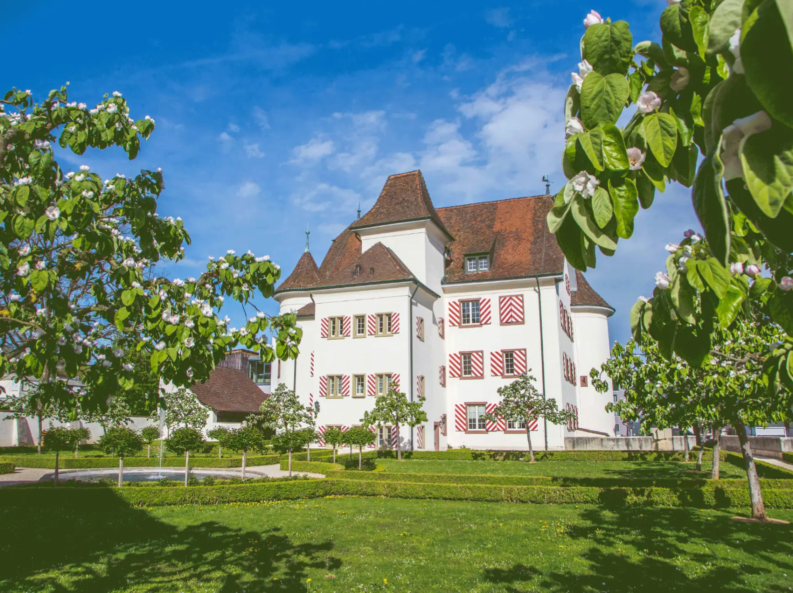 Château d'Aesch entouré d'arbres fruitiers et d'un espace vert soigné sous un ciel bleu.