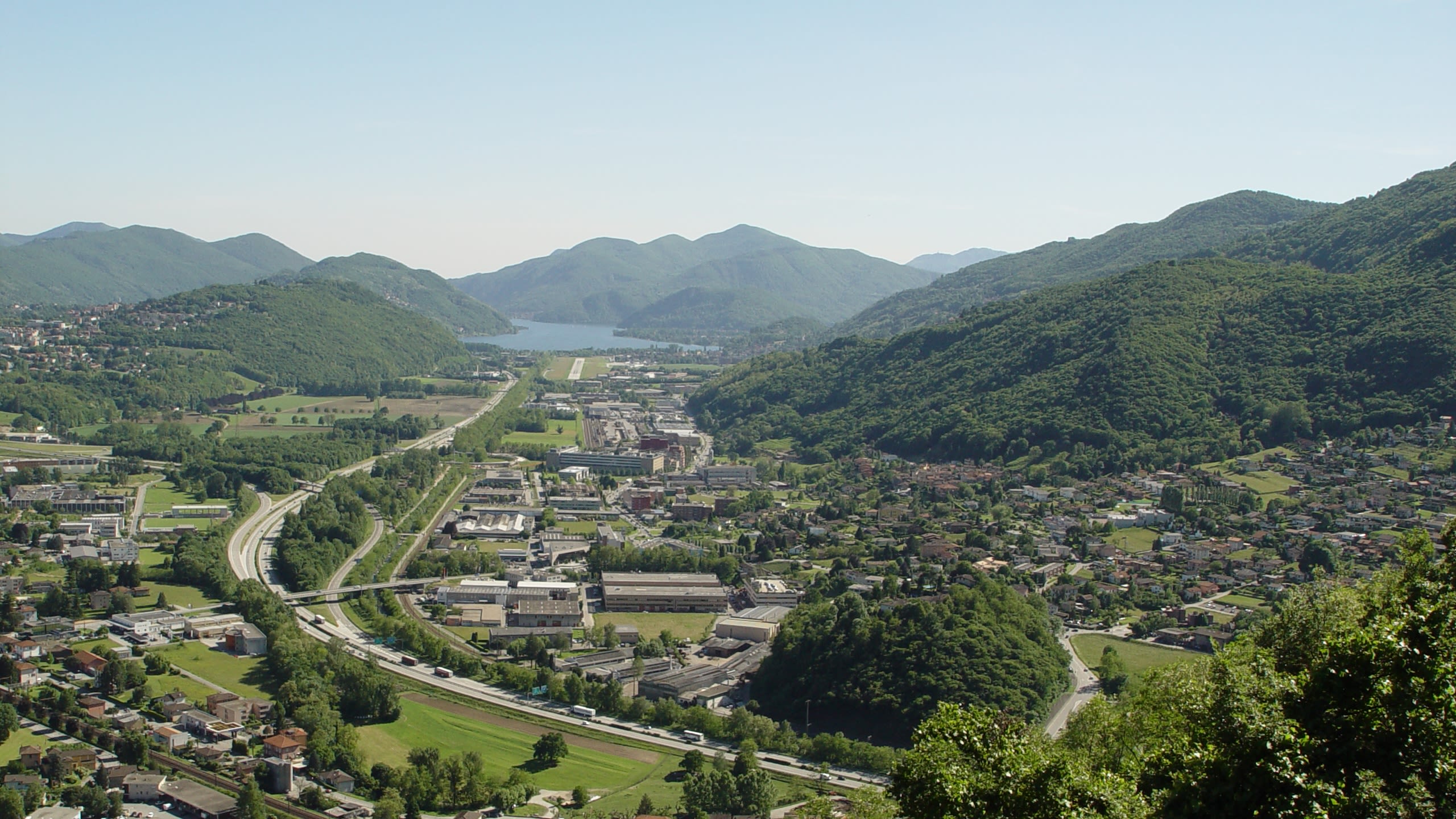 Vallée verte au Tessin avec des collines, des maisons, des routes et un lac au fond.