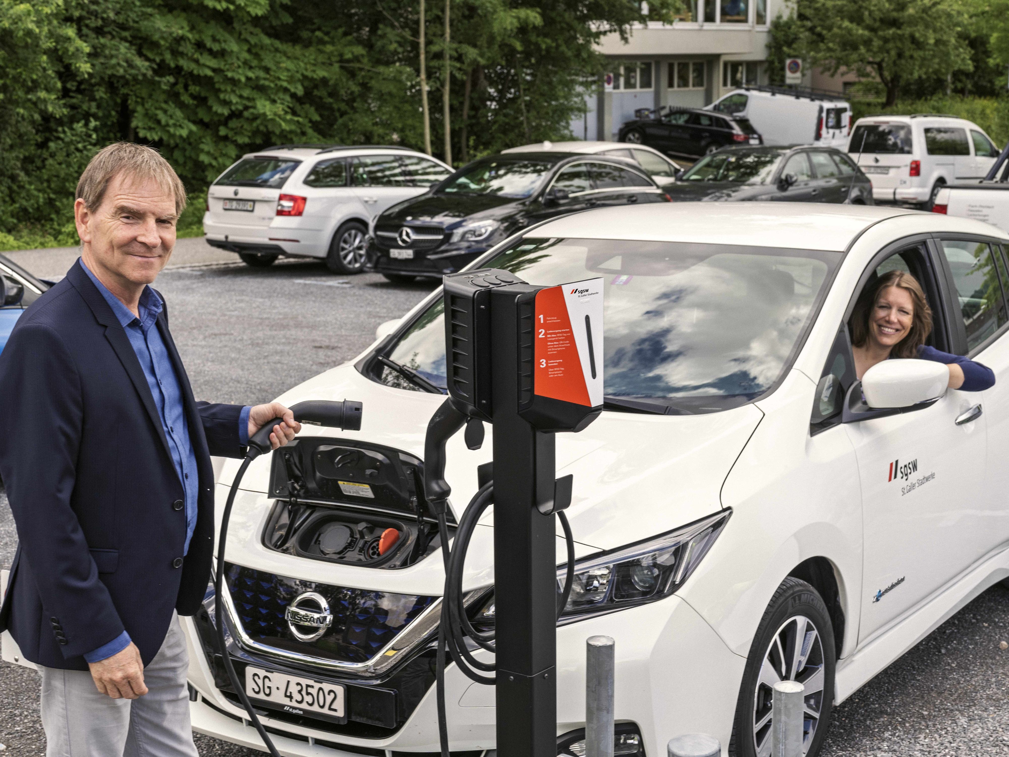 Un homme connecte la voiture électrique à la borne de recharge. Une femme est assise dans la voiture et regarde par la fenêtre ouverte.