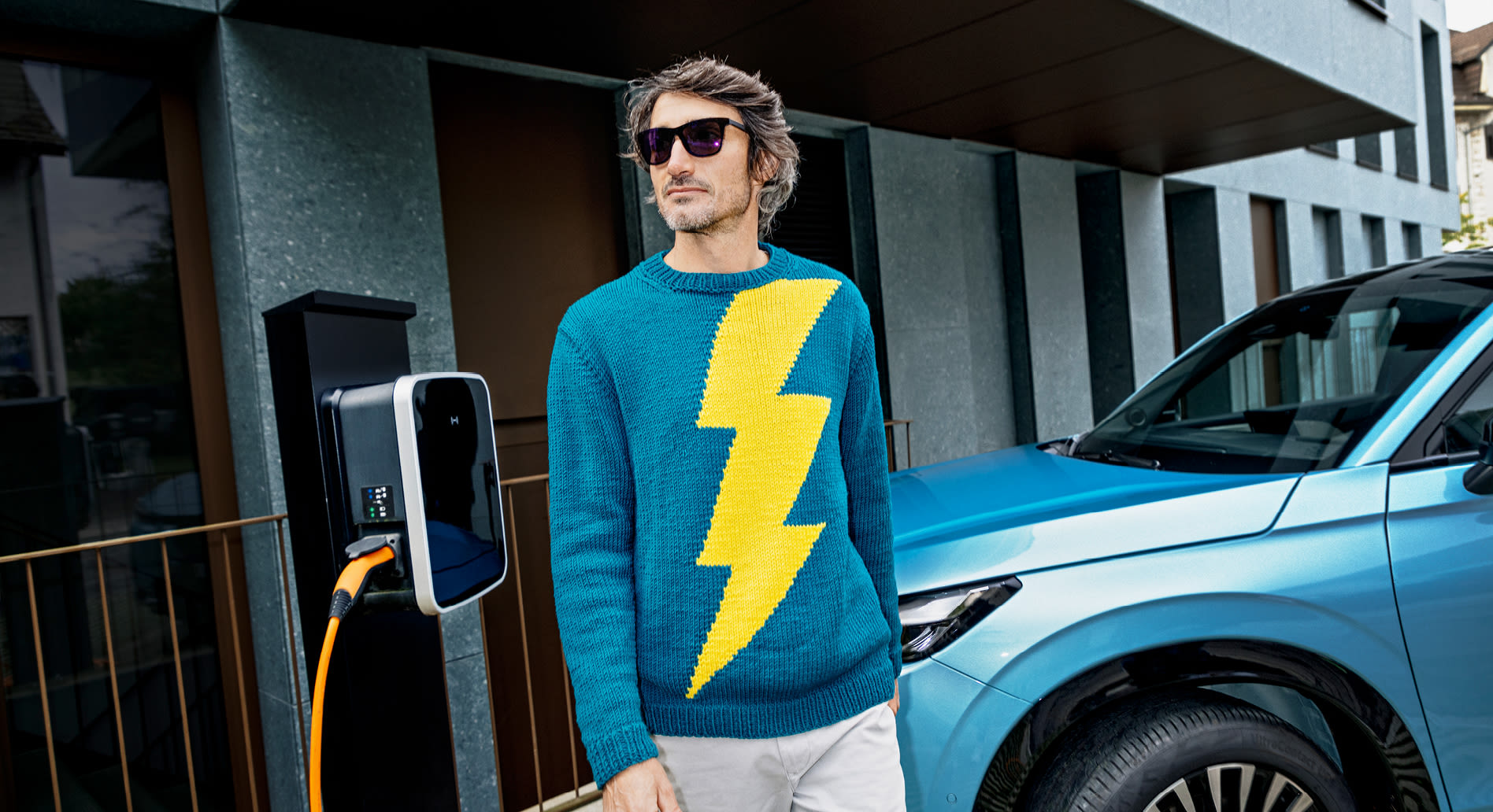 Un uomo con un maglione blu elettrico decorato con un fulmine si trova davanti a un'auto in carica