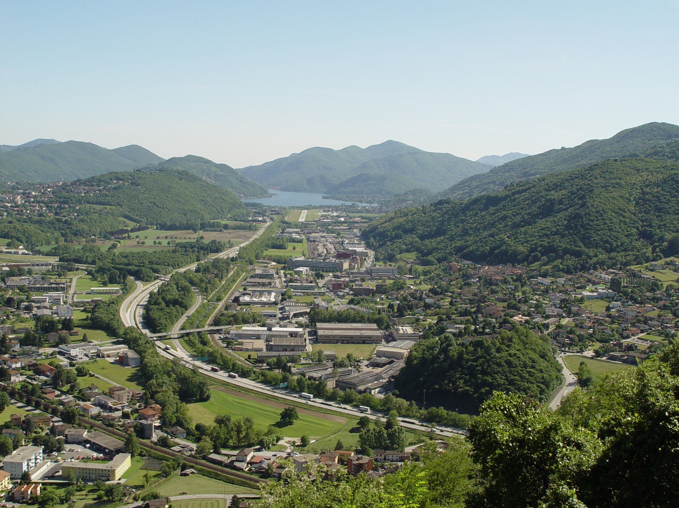 Vallée verte au Tessin avec des collines, des maisons, des routes et un lac au fond.
