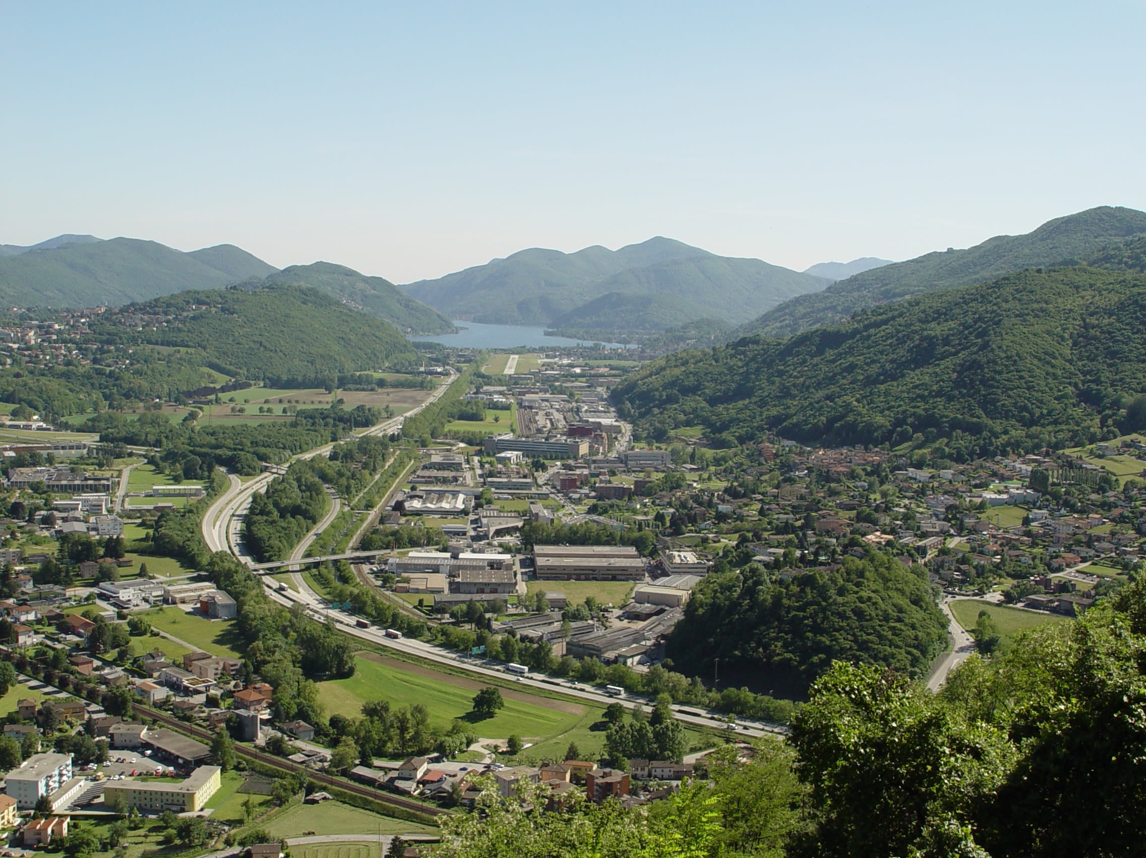 Vallée verte au Tessin avec des collines, des maisons, des routes et un lac au fond.