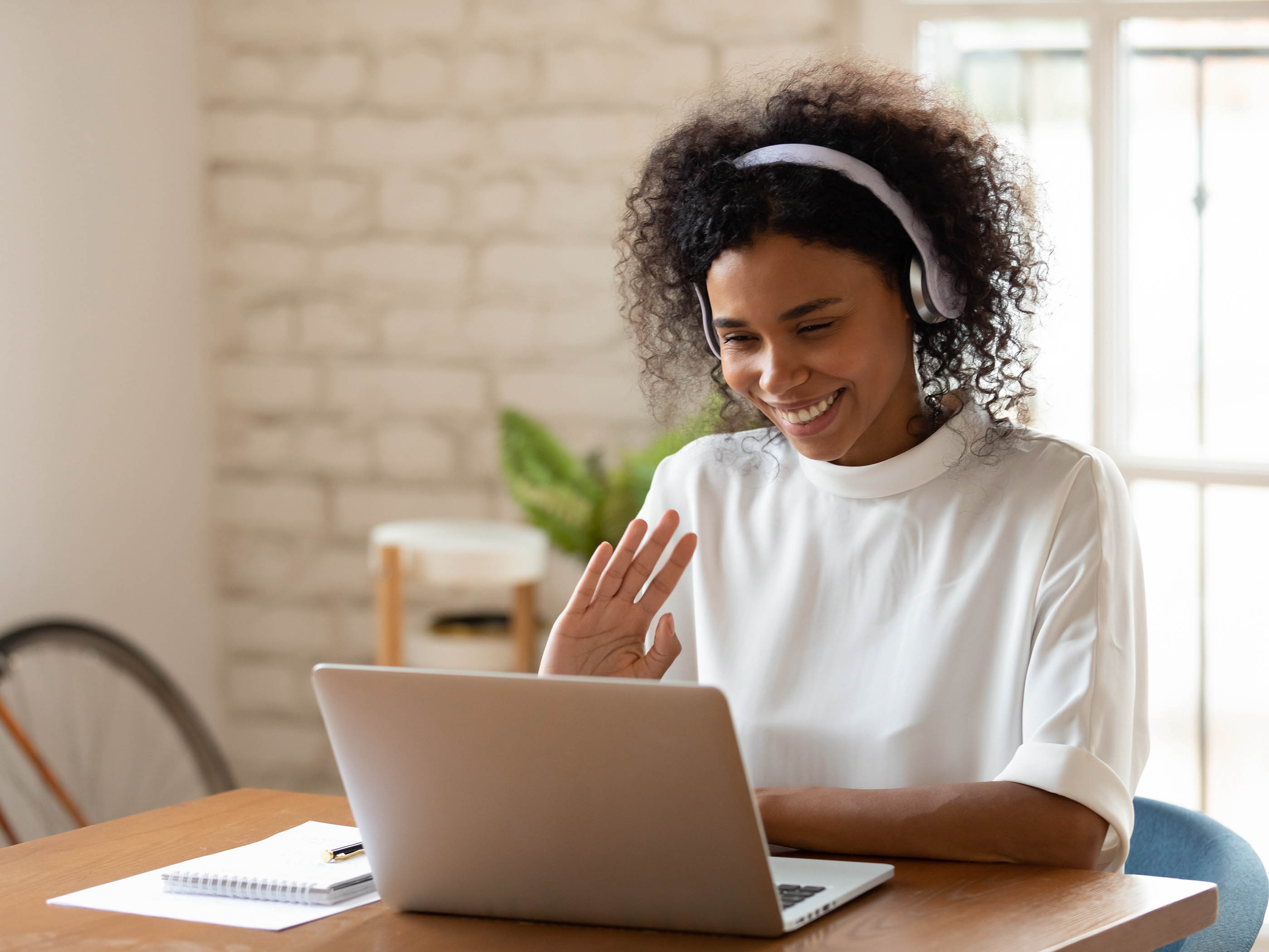 Une femme est assise à une table avec un casque et fait signe de la main à son ordinateur portable.