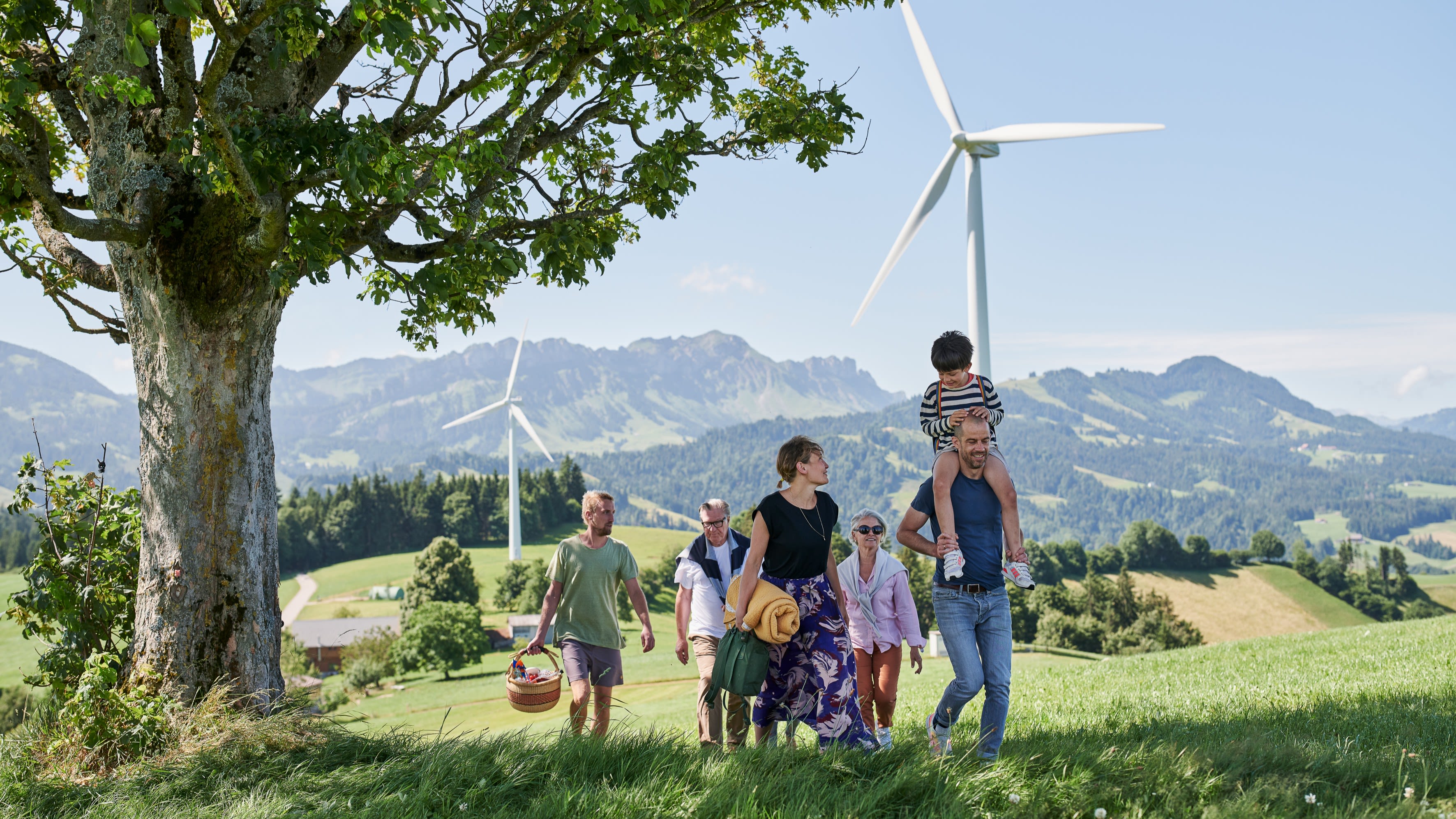 Una famiglia con bambini in gita. Hanno portato un cestino da picnic. Sullo sfondo si vedono le montagne e le pale eoliche.