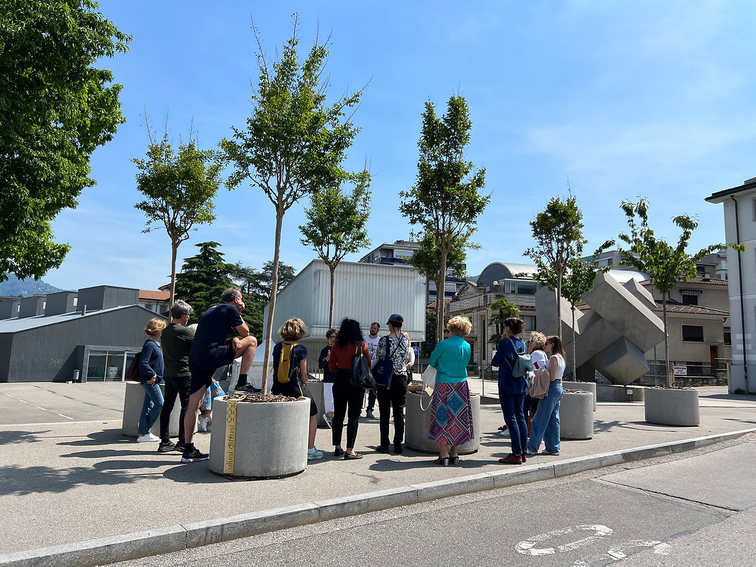 Menschen stehen auf einem Platz in Chiasso zwischen Bäumen in Töpfen.