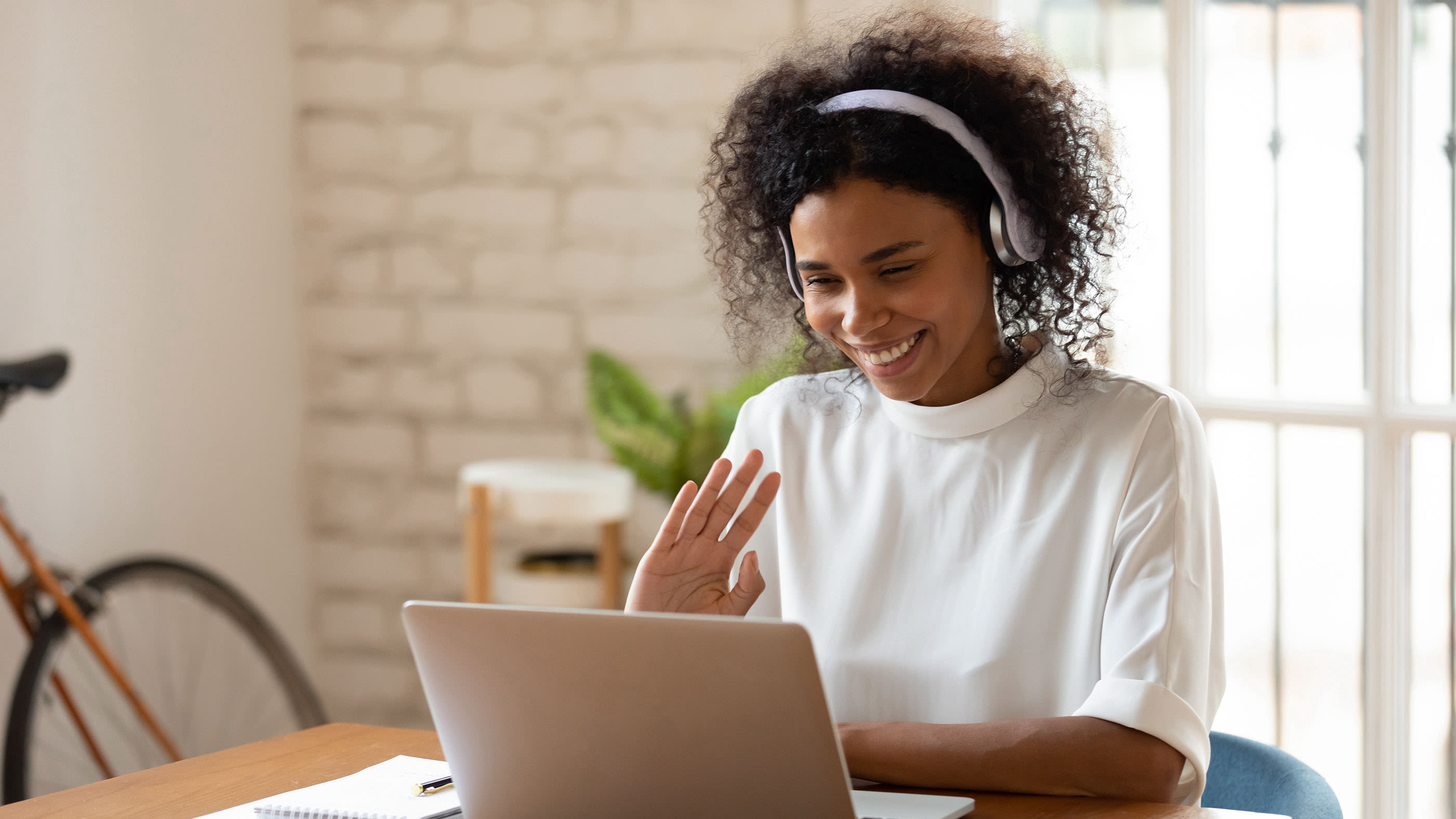 Une femme est assise à une table avec un casque et fait signe de la main à son ordinateur portable.