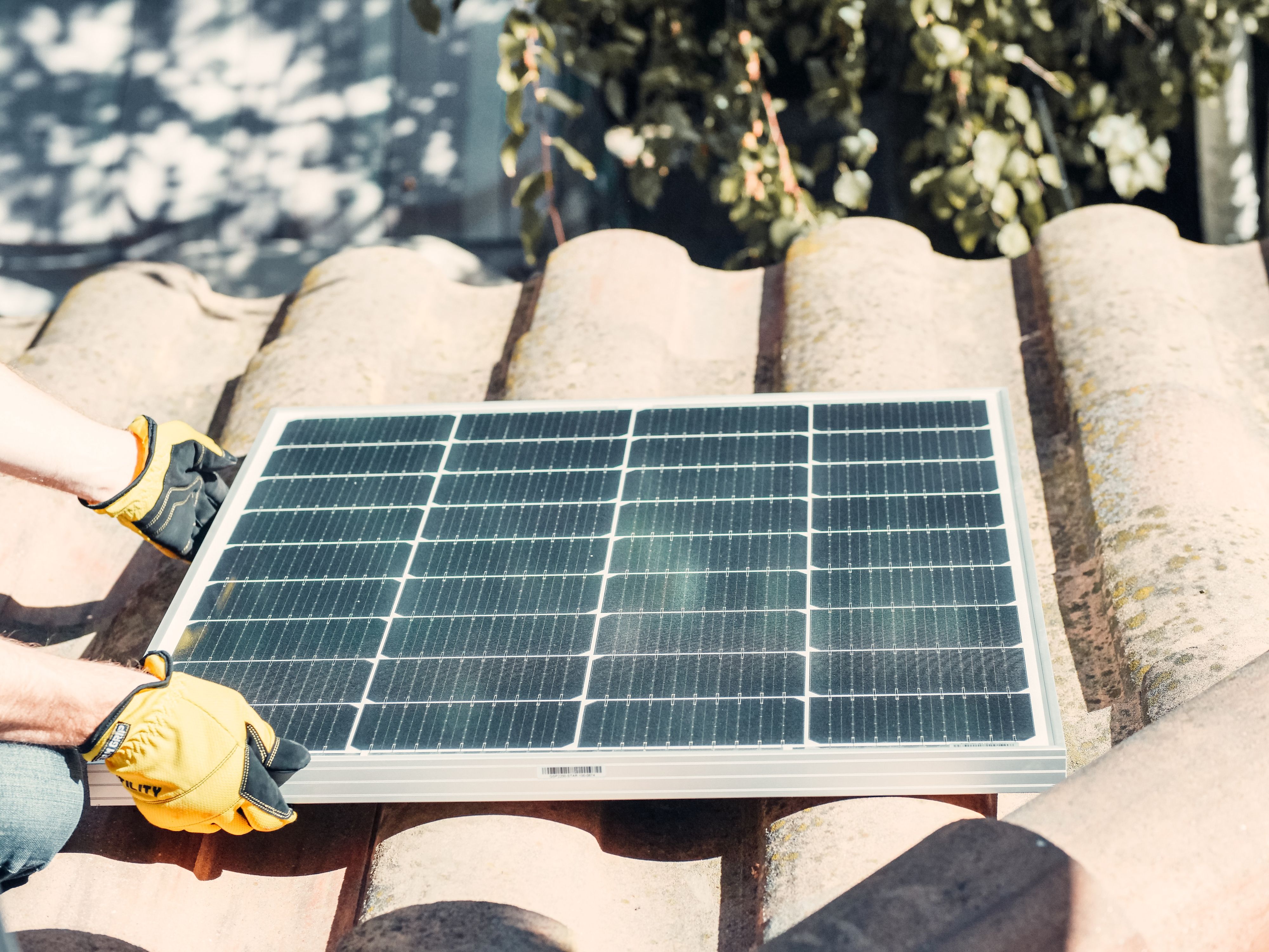 Zwei behandschuhte Hände halten einen Solarpanel auf einem Haus mit Ziegeldach.