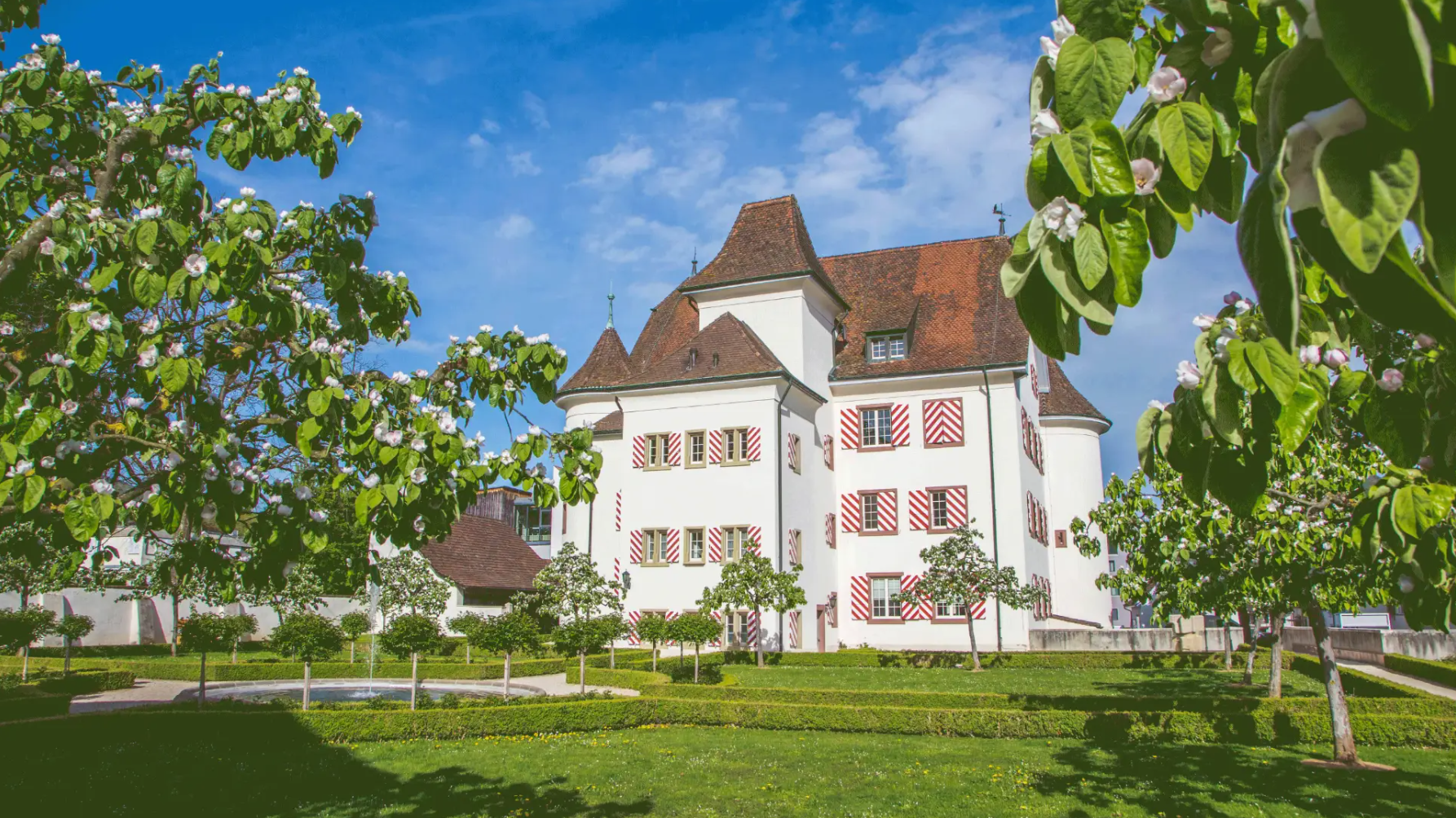 Castello di Aesch circondato da alberi da frutto e un curato parco verde sotto un cielo azzurro.
