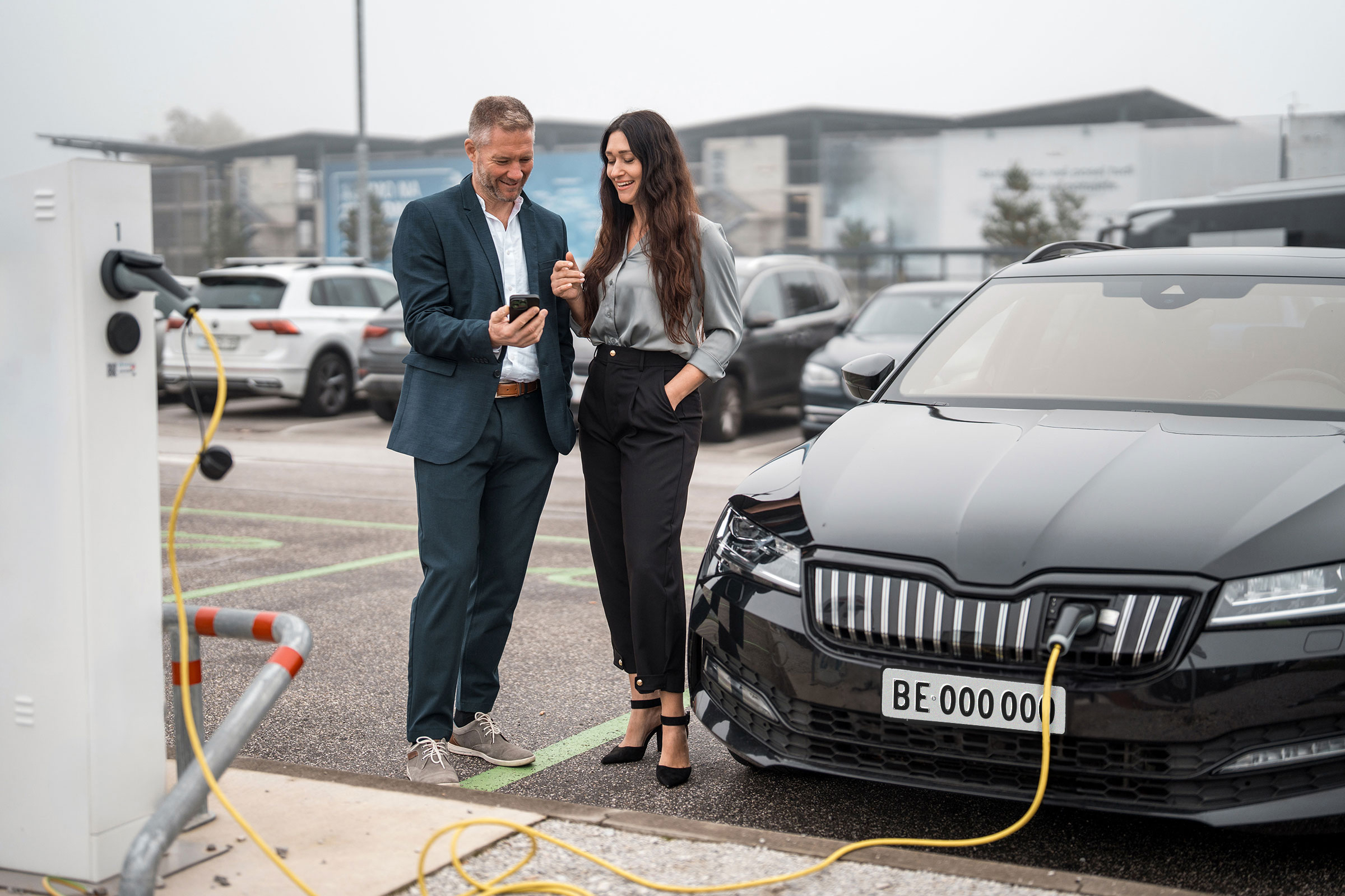 Due persone sono accanto a un’auto elettrica in ricarica e osservano insieme uno smartphone.