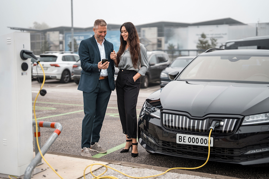 Due persone sono accanto a un’auto elettrica in ricarica e osservano insieme uno smartphone.