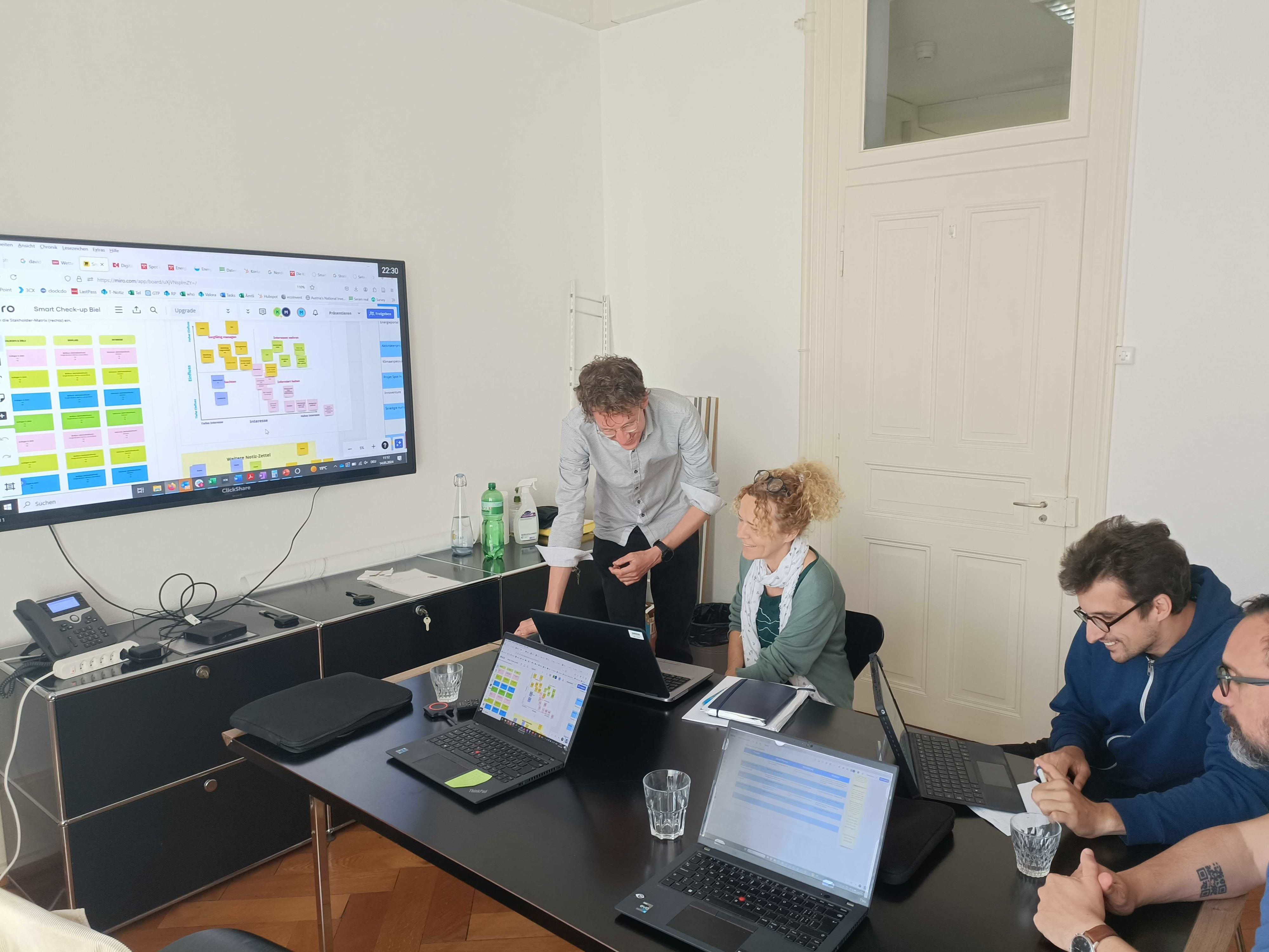 Une salle de réunion avec quatre personnes autour d'une table de conférence, travaillant sur des ordinateurs portables et discutant. Des diagrammes et des notes sont visibles sur un écran accroché au mur.