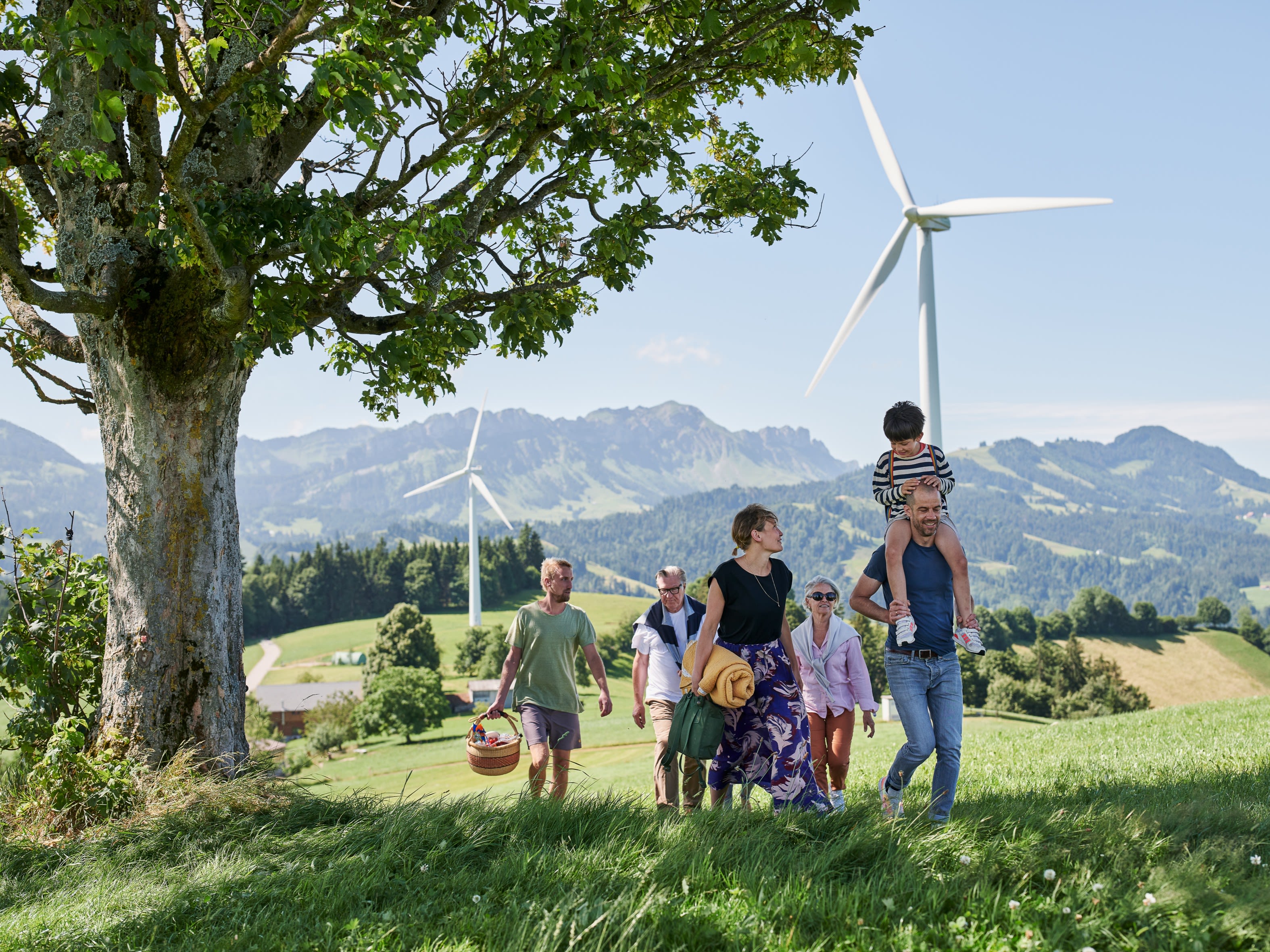 Una famiglia con bambini in gita. Hanno portato un cestino da picnic. Sullo sfondo si vedono le montagne e le pale eoliche.
