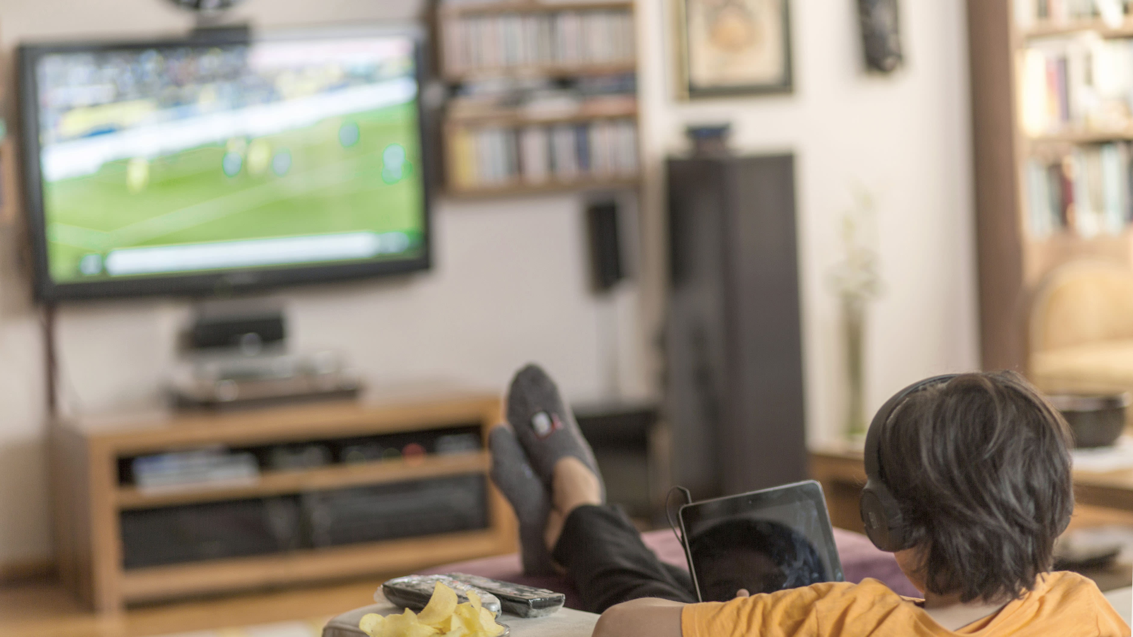 Junge mit Kopfhörern liegt auf dem Sofa und schaut Fussball im Fernseher.