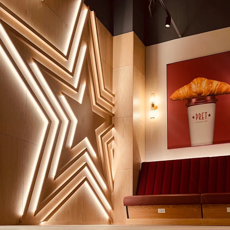 The Pret-A-Manger star adorns a brown wall, with a picture of a croissant on a coffee cup on another wall. A red sofa is underneath the picture.