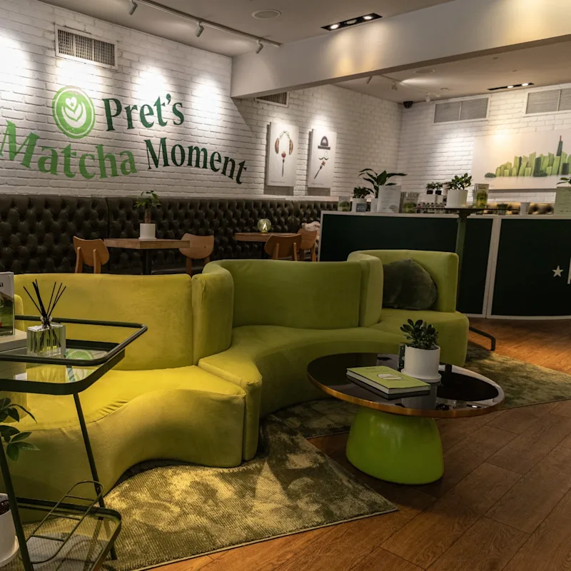 A coffee shop with green sofa's and a green coffee table. A white wall is in the background with text that reads Pret's Matcha Moment