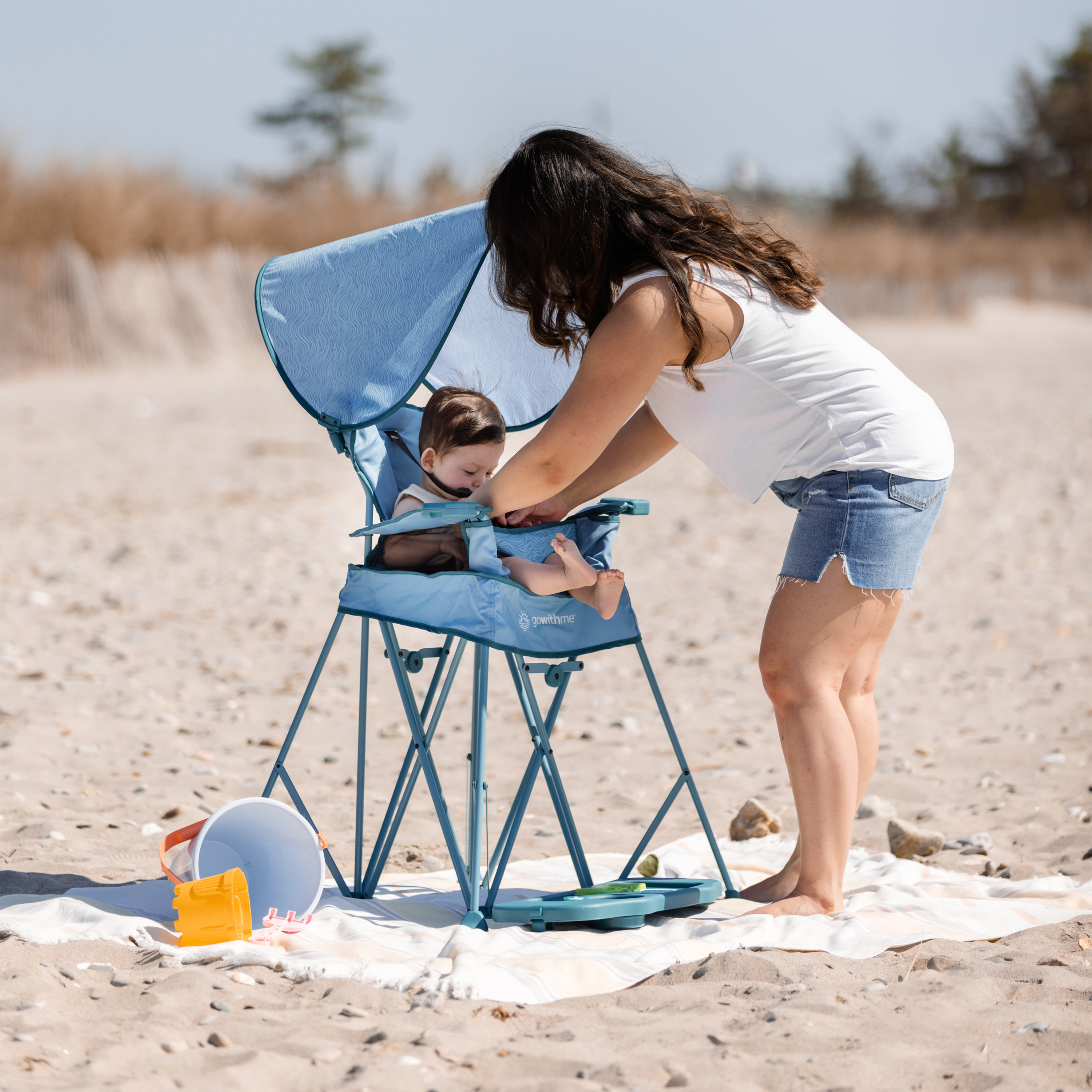 Woman using a blue and white portable high chair with a baby on the beach.