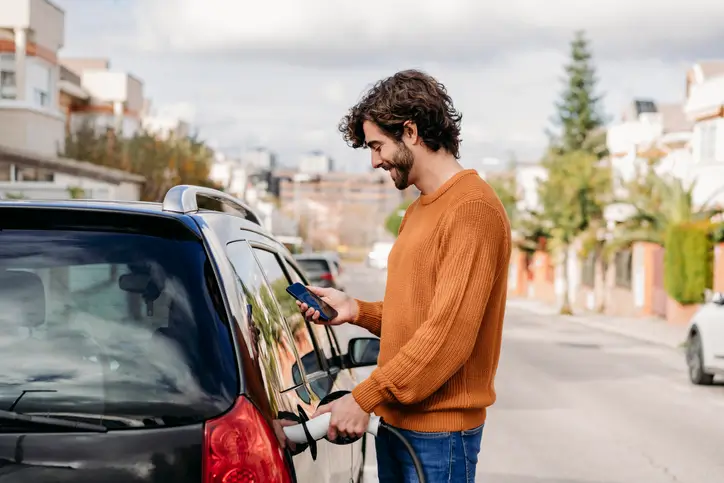 A photo of a young male in an orange jumping standing by his car as he charges it. He is smiling and looking down at his phone