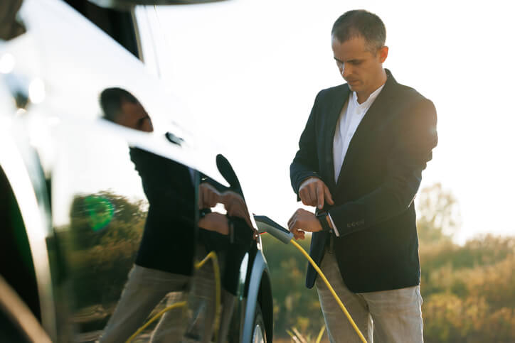 A photo of a man charging his electric car whilst he looks at his watch