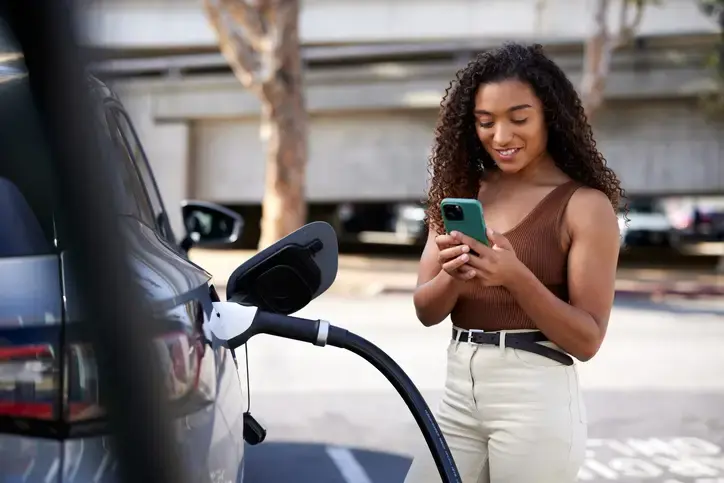 A photo of a smiling young woman using her mobile phone by her car as it gets charged at the station