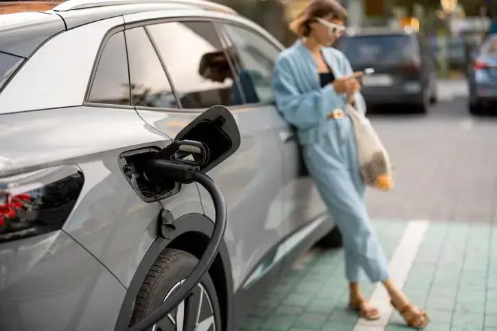 A photo of a stylish woman standing in the background on her phone leaning against her electric car. In the foreground there is a close up of her cars port with a charging cable plugged into it.