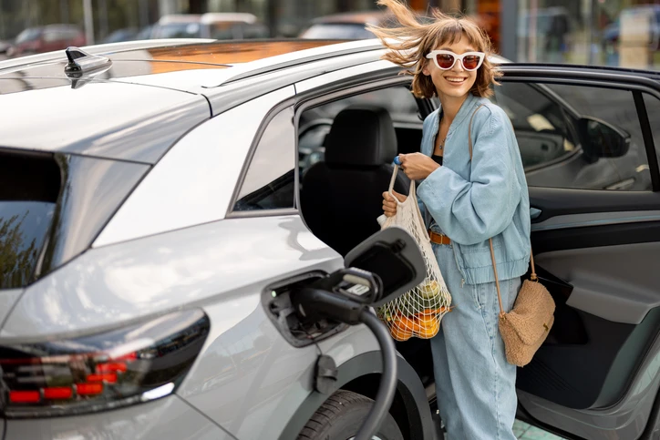 A photo of a stylish young woman wearing sunglasses and a denim co-ord. She is charging her electric car whilst she puts groceries in the back seat