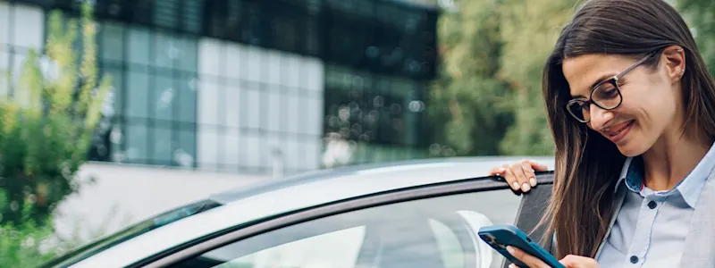 Frau steht am Auto uns schaut nach News der Contipark auf ihrem Smartphone