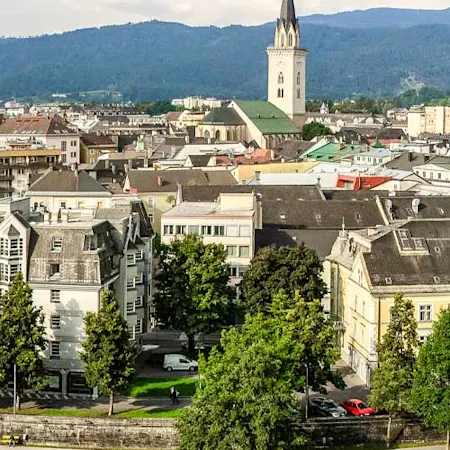 Parken in Villach : Auf einem Panoramabild ist die Stadt Villach zu sehen. Im Zentrum der Stadt erhebt sich der Stadtpfarrturm, der alle anderen Gebäude überragt, während die Drau die Stadt umfließt.