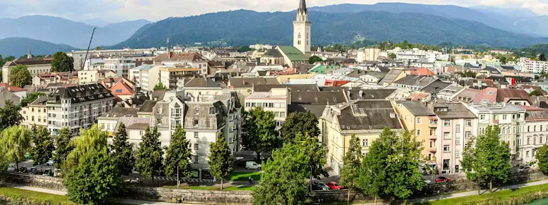 Parken in Villach : Auf einem Panoramabild ist die Stadt Villach zu sehen. Im Zentrum der Stadt erhebt sich der Stadtpfarrturm, der alle anderen Gebäude überragt, während die Drau die Stadt umfließt.