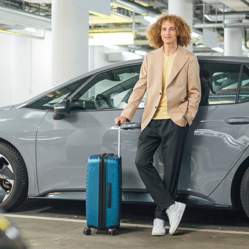 junger Mann lehnt mit überkreuzten Beinen und Koffer lässig an einem parkenden Auto in der Tiefgarage