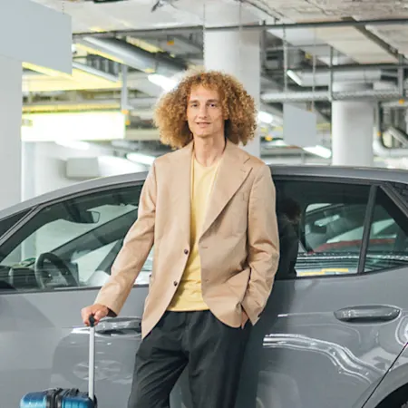 junger Mann lehnt mit überkreuzten Beinen und Koffer lässig an einem parkenden Auto in der Tiefgarage